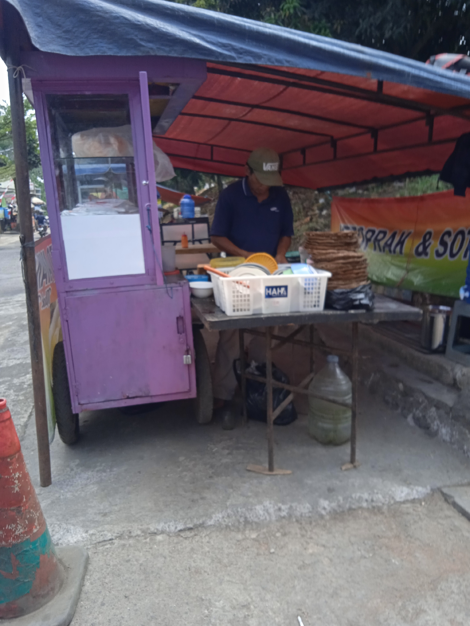 A person wearing a dark blue polo shirt and a light brown cap is standing behind a small food cart. The cart is primarily purple with a clear glass front display. The person appears to be preparing or serving food, with their hands positioned over a table laden with various items. A stack of flat, round food items, possibly crackers or bread, is prominently displayed on the table, along with a white plastic basket containing more food items, a bowl, and a yellow fan-shaped object. To the right of the table, a large clear plastic water jug is visible, as well as a metal canister. In the background, a banner with orange and green lettering, possibly advertising food items, is partially visible. The scene is set outdoors, on a concrete surface, with a red and green traffic cone in the lower left foreground. The environment suggests a street-side or market setting in Cileunyi, Indonesia.