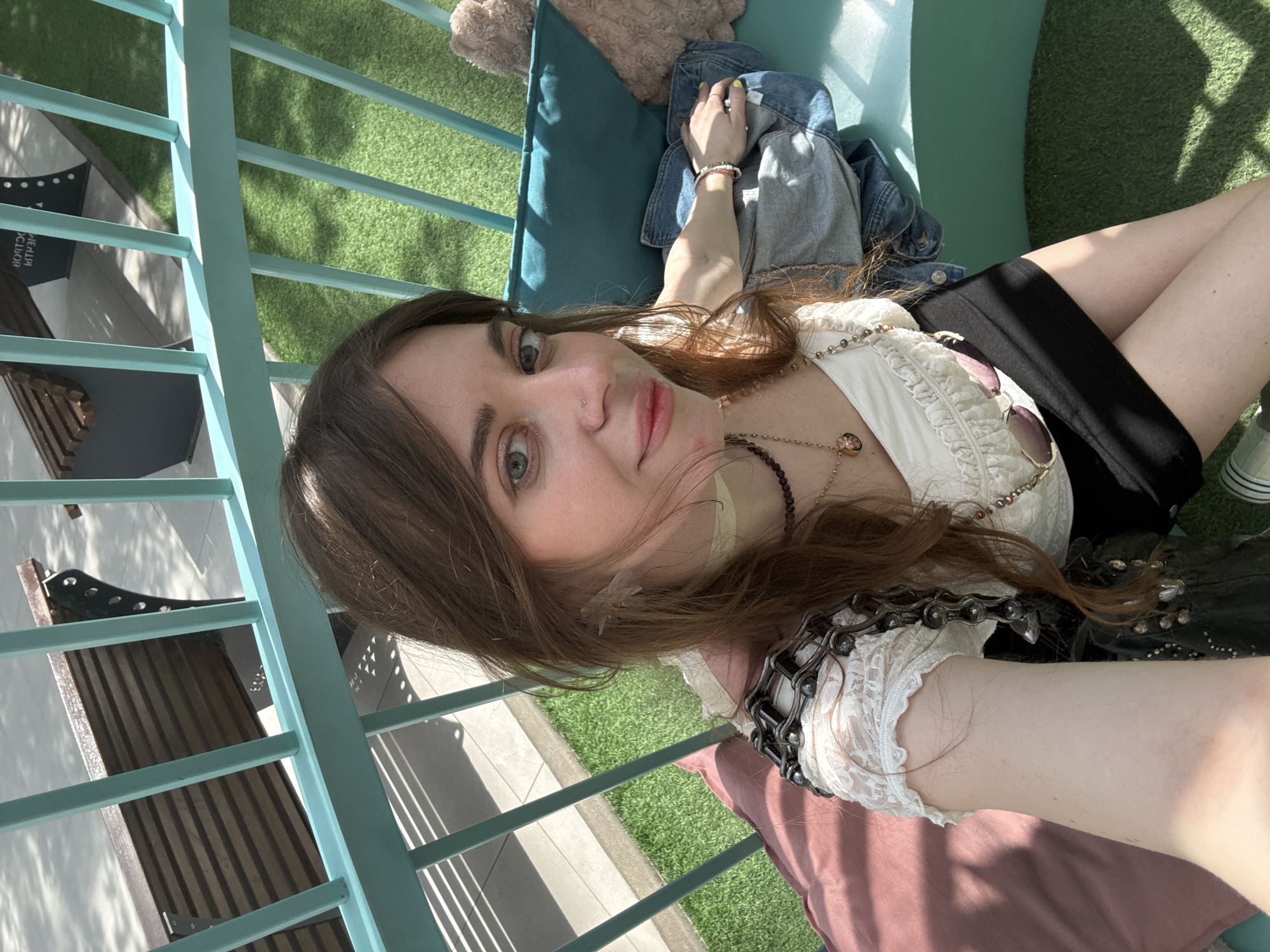 A young woman is lying on a bench in a public outdoor area. The bench is made of light blue metal and has a green cushion and a pink pillow. There are patches of green grass in the background, and another bench on the left. The woman is wearing a white top, a black skirt, and necklaces. She is lying on her side, looking at the camera. A denim jacket is draped on the bench nearby. The location is Moscow, Russia.