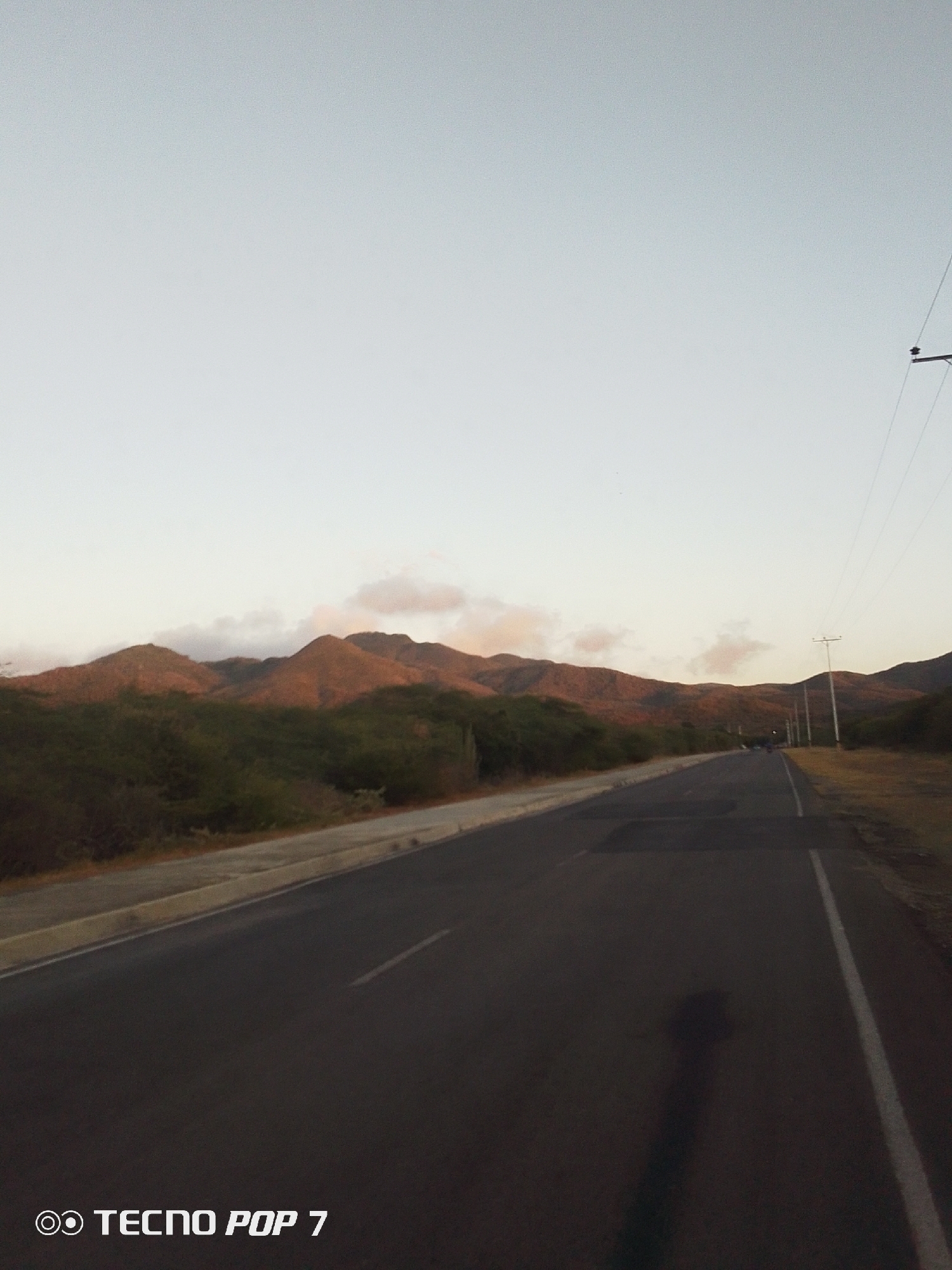 A long, asphalt road stretches into the distance, framed by mountains in the background under a pale sky. The road has a white line on its right side and a concrete curb on the left, beyond which is low green vegetation. Power lines and poles run along the right side of the road. The image was taken on a TECNO POP 7. The road is likely located near Altagracia, Venezuela, given the mountainous terrain and sparse vegetation.