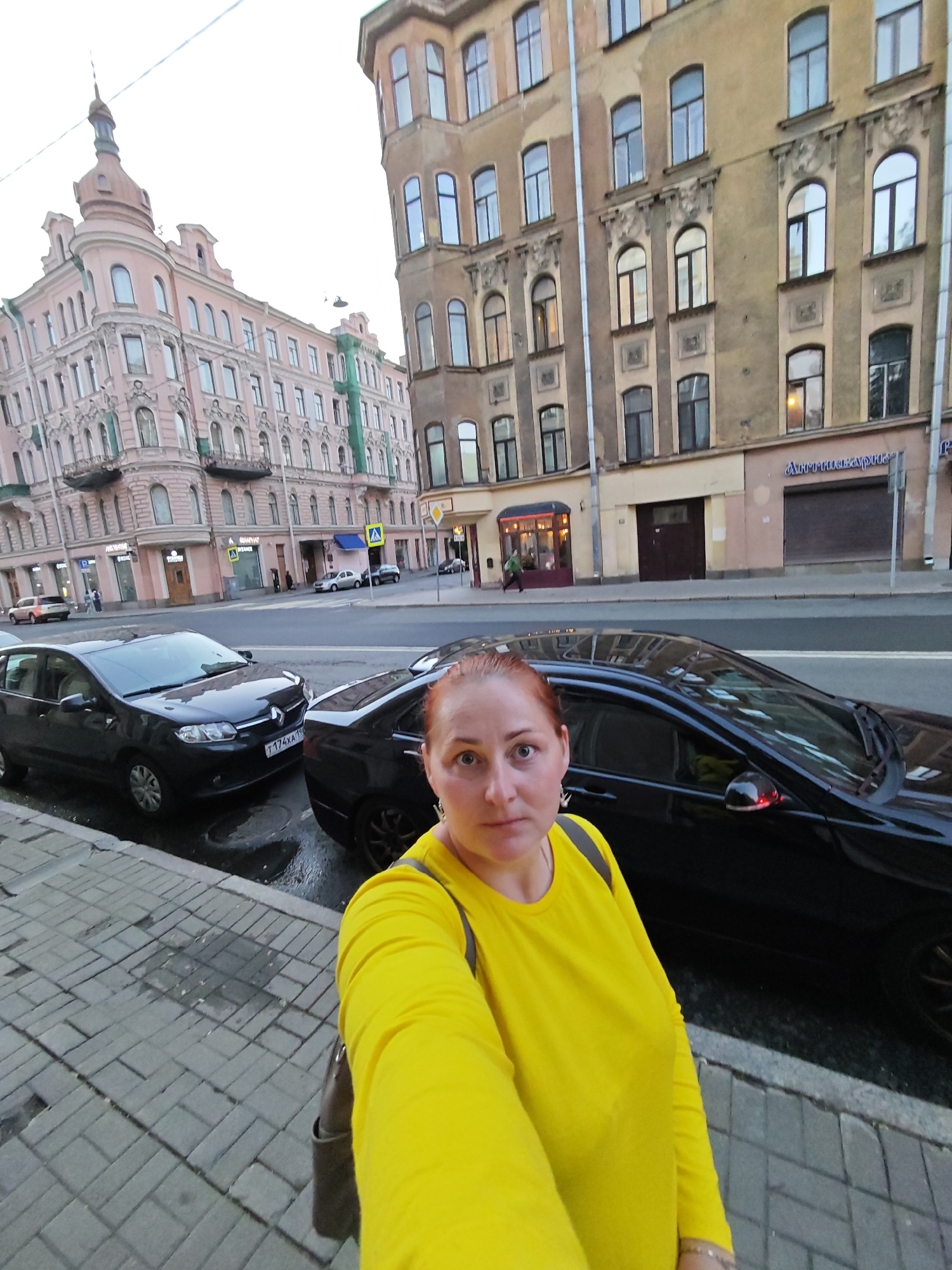 A medium shot is captured from a low angle, looking up towards a woman with red hair and a yellow shirt, taking a selfie in Saint Petersburg, Russia. The woman is positioned on a sidewalk next to a street with parked cars. Behind her, buildings with ornate architecture line the street, with a pink building on the left and a beige building on the right. A black car is parked in front of the woman, and another black car is visible further down the street. The sky is overcast. The scene appears to be on a city street.