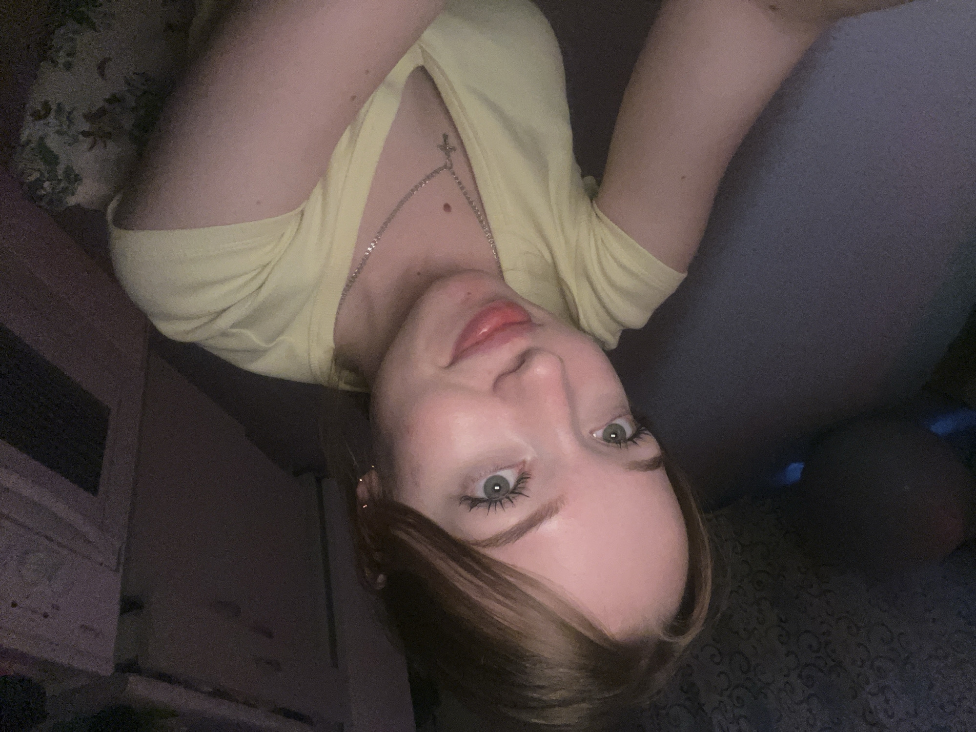 A young woman is lying on a bed, wearing a yellow v-neck shirt and a silver necklace with a cross pendant. She has fair skin, light eyes, and brown hair. Her head is tilted to the side, and she appears to be looking directly at the camera. The background includes part of the bed, a wall with a patterned wallpaper, and a piece of furniture, likely a dresser or cabinet. There is no information to indicate the location is in the city of Chaikovsky, Russia.
