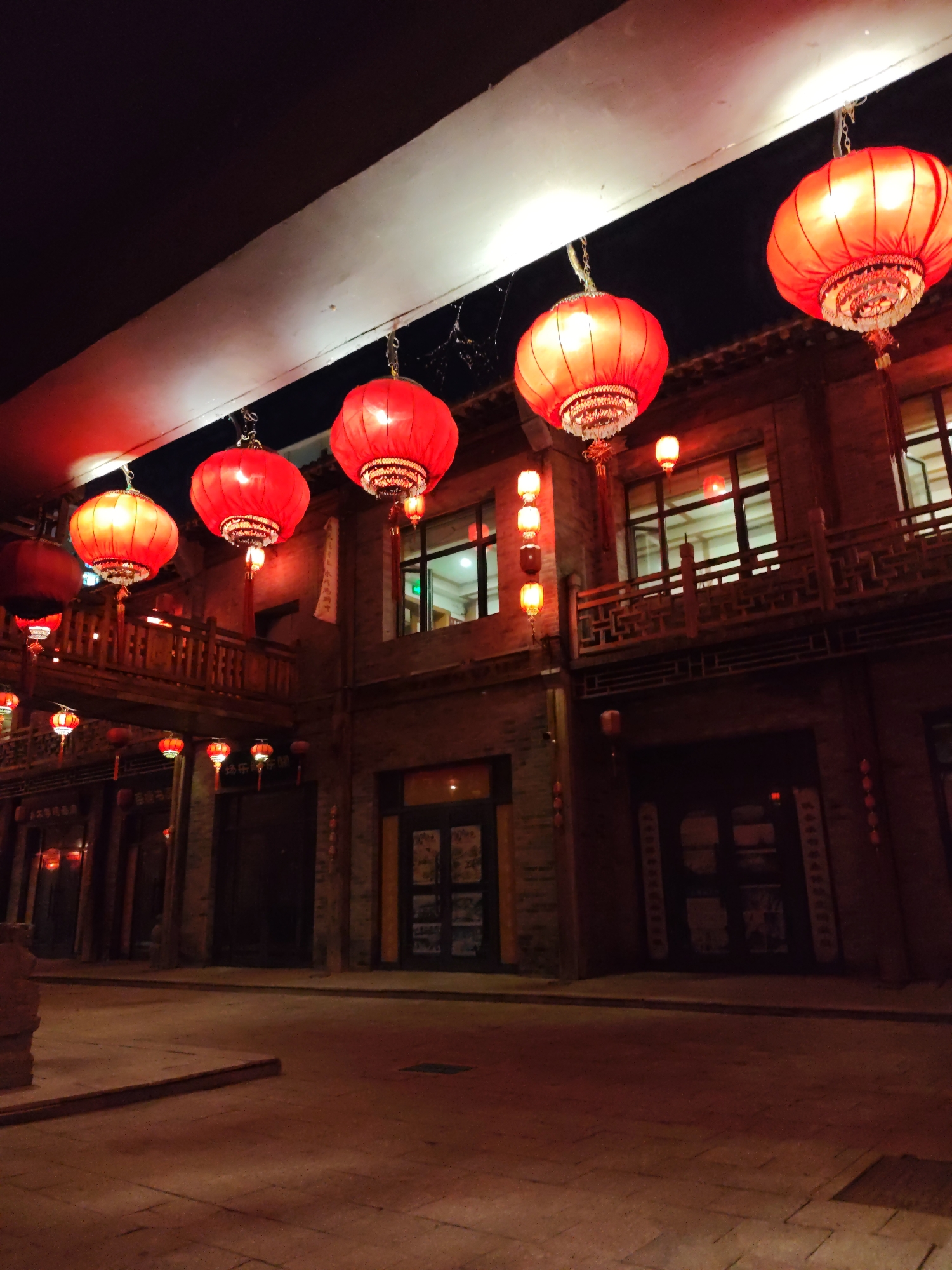 The image captures a nighttime scene in Lindian, China, featuring a street lined with traditional brick buildings adorned with red lanterns. The lanterns, illuminated with a warm glow, hang from wires stretched across the street, creating a festive and atmospheric ambiance. The buildings appear to be commercial establishments, with visible windows and doorways. A wooden walkway or bridge can be seen spanning between some of the buildings on the left side of the image. The cobblestone street is wet in places, reflecting the light of the lanterns. The overall scene suggests a lively cultural or festival setting.