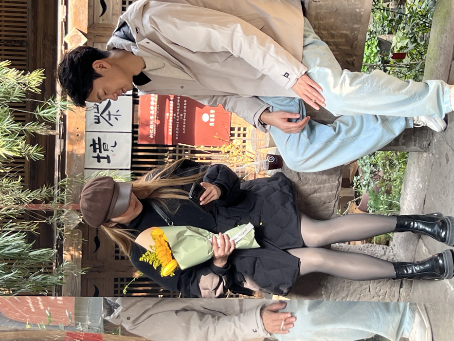 A man and woman are seated outdoors. The man wears a light jacket and light blue jeans. The woman wears a brown cap, a black jacket, a black skirt, and black boots. She holds a bouquet of yellow flowers wrapped in green paper. They appear to be on a stone ledge or bench. The background includes plants, traditional-style buildings with Chinese characters on signs, and other people.
