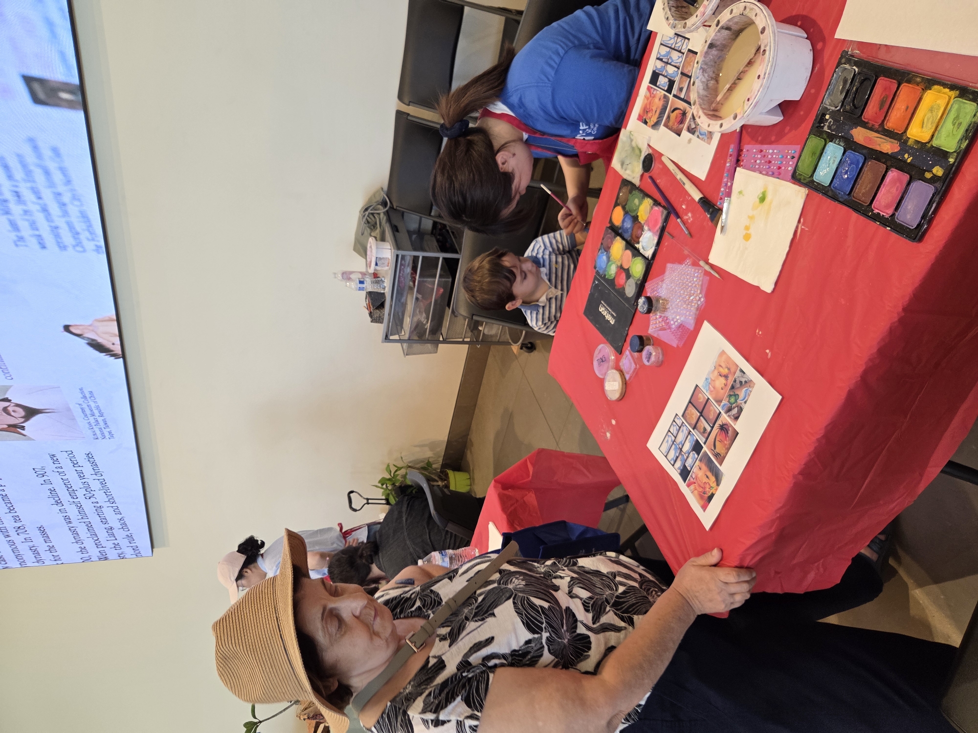 A woman wearing a wide-brimmed straw hat and a black and white floral print shirt is standing at a table covered with a red tablecloth. Her gaze is directed downward at the table. Next to her, a child in a striped shirt is seated. A woman in a blue shirt with a red apron is painting the child's face. On the table, there are various art supplies including paint palettes, brushes, a container of water, and a printed sheet with images. In the background, there are chairs, a trolley with more supplies, and a large screen displaying text and an image. The environment appears to be an indoor public space, possibly a community center or an event.