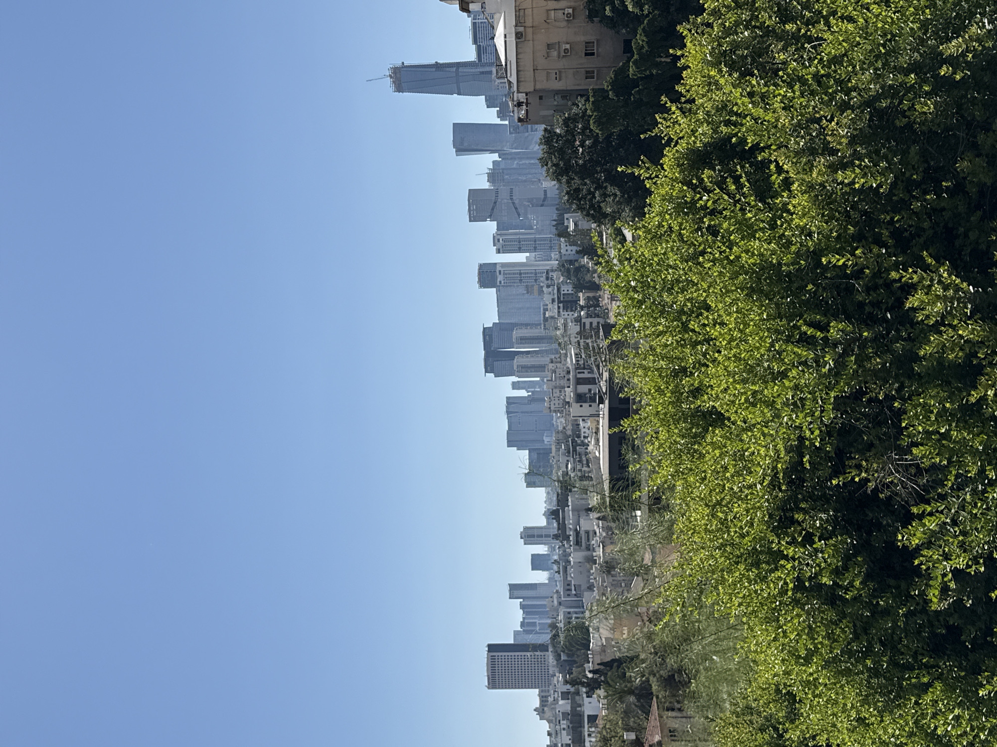 The image shows a cityscape in Givatayim, Israel. A row of buildings, including high-rises, stretches across the middle of the frame. Green trees partially obscure the buildings on the right side. A clear blue sky takes up the left side of the image.