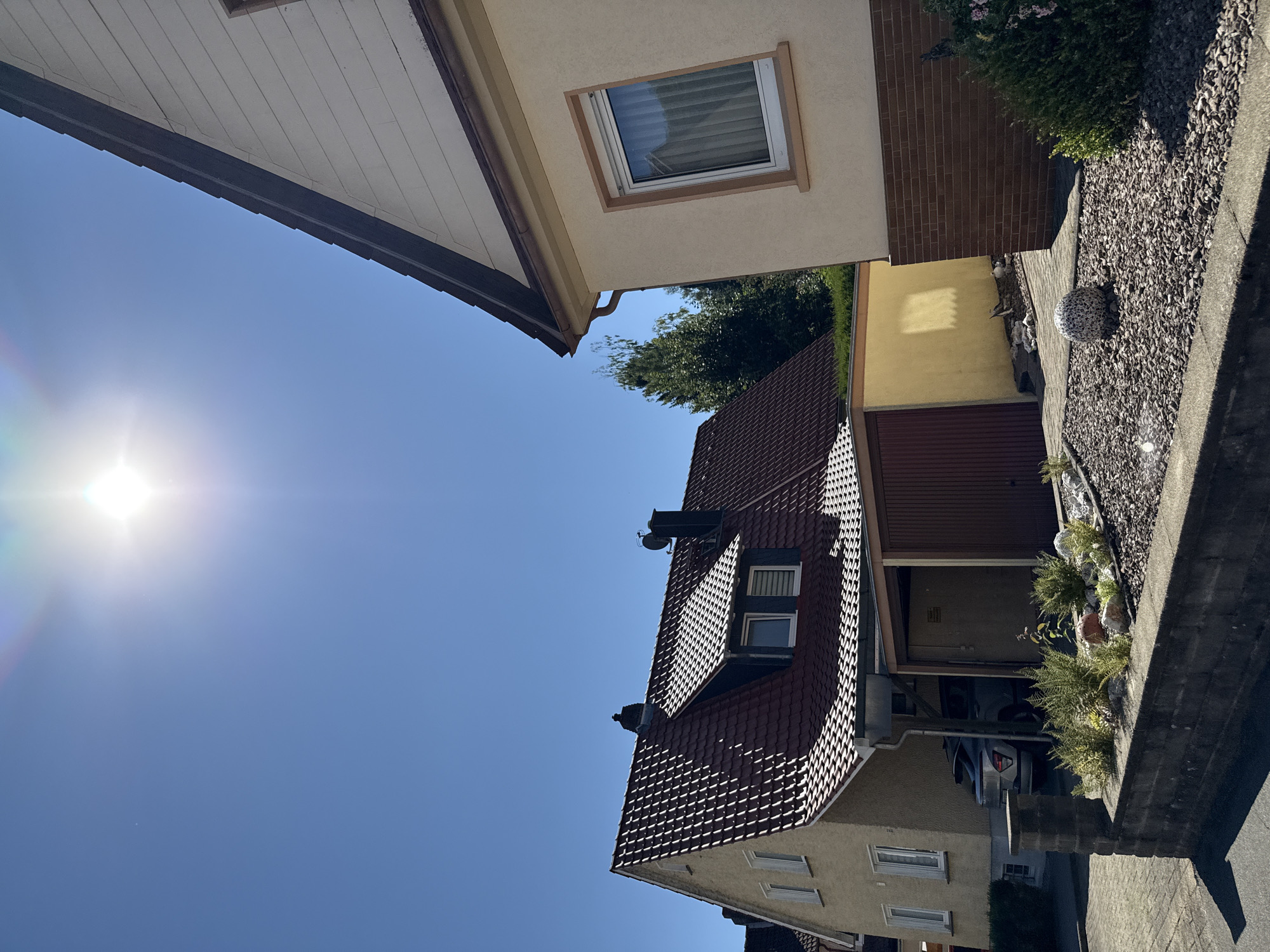 The image depicts a street scene in Bad Lauterberg im Harz, Germany, on a bright, sunny day. The foreground features a low stone wall bordering a garden area covered with small, dark gray pebbles and a few larger decorative stones. Several small green plants are visible in the garden, including ferns. To the right of the garden, a portion of a light-colored house is visible, with a window featuring light-colored curtains and a brown brick base. A dark green evergreen bush is situated in front of this house.
In the mid-ground, a two-story house with a light yellow facade and a red tiled roof is prominent. This house has several windows on its upper floor and a dormer window with two panes on the roof. Adjacent to this house is a covered carport with a dark brown garage door. A silver SUV is parked under the carport. Behind the carport, a single-story structure with a yellow wall is visible.
The background includes a clear blue sky with the sun prominently positioned near the top center, creating a lens flare effect. Tall green trees are visible behind the houses. The houses appear to be residential properties. No people are visible in the image. The scene is well-lit due to the strong sunlight.