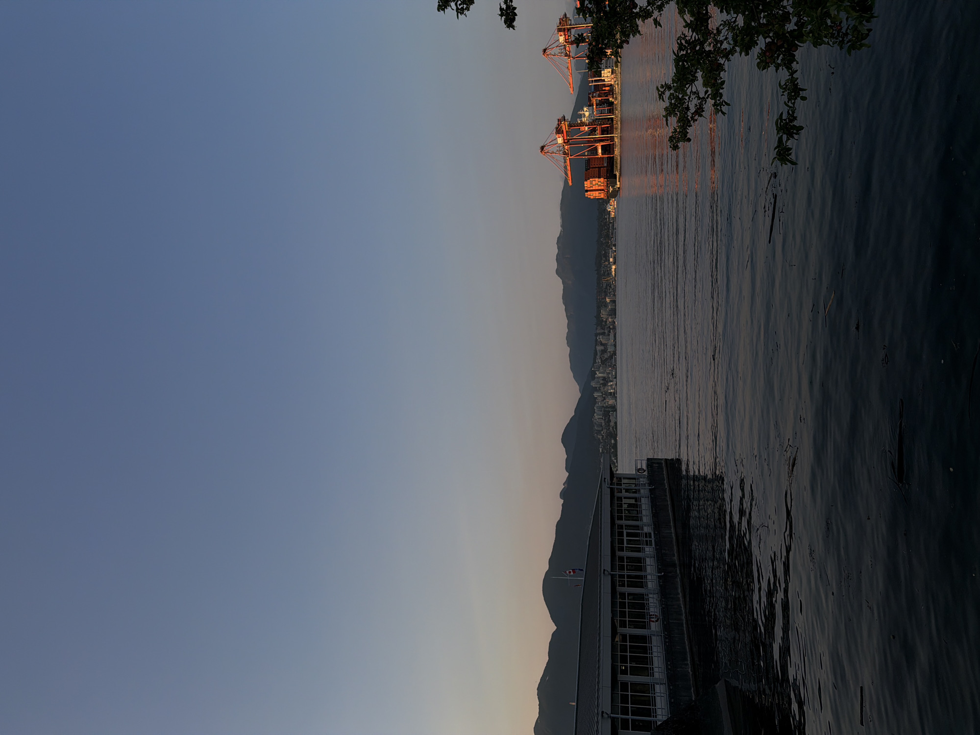 The image depicts a serene waterside scene in Vancouver, Canada, likely at dusk given the warm glow on the distant mountains and the muted sky. In the foreground, a dark, calm body of water stretches towards the horizon, with a few pieces of debris floating on its surface. To the left, a long, modern building with a flat roof and numerous windows runs parallel to the water, its reflection faintly visible in the water. A flag is visible atop the building.
Further out in the water, on the right side of the frame, a large cargo ship is docked, dwarfed by two massive, bright red gantry cranes. These cranes are positioned over the ship, suggesting active cargo operations, though no visible human activity is present. The ship itself is laden with shipping containers.
In the distance, a range of dark, silhouetted mountains forms the backdrop, with a city skyline visible in the gap between the mountains and the water. The sky is a gradient of pale blue at the top, transitioning to a soft, warm hue near the horizon. The overall atmosphere is one of tranquility, with the industrial presence of the port juxtaposed against the natural beauty of the mountains and water.
