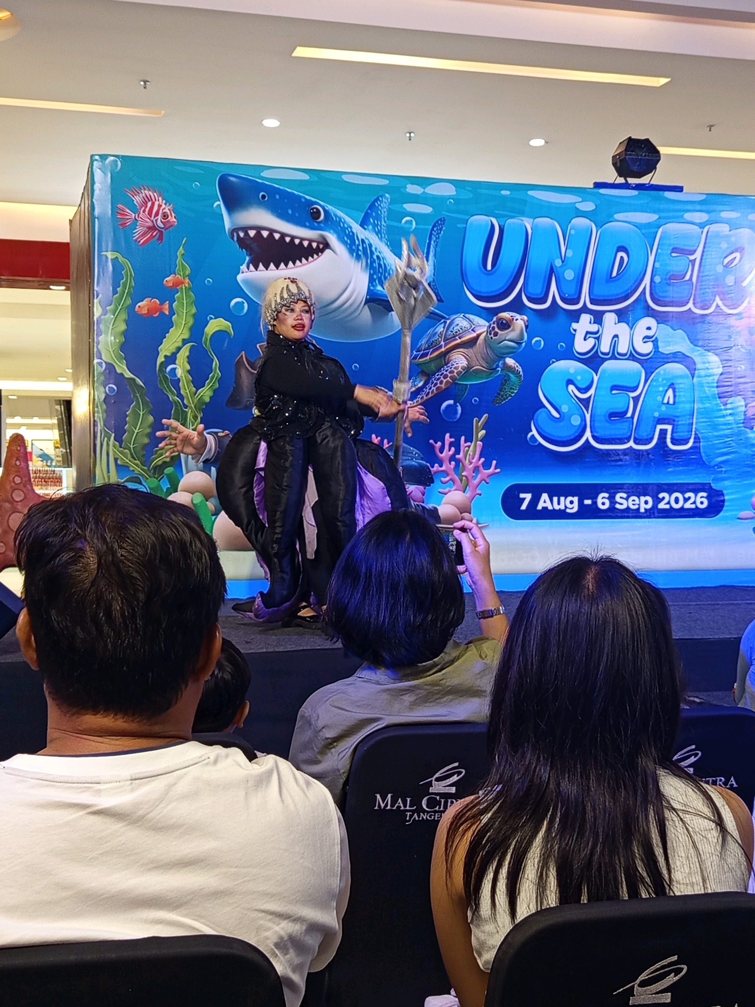 In an indoor venue in Ciakar, Indonesia, a performer dressed as an undersea character stands on a stage in front of a large backdrop. The backdrop features a blue, watery scene with images of a shark, a turtle, fish, seaweed, and coral. Text on the backdrop reads "UNDER the SEA" and "7 Aug - 6 Sep 2026." The performer wears a black sequined top, black pants, and a purple, tentacle-like skirt. They have a dramatic makeup style with elaborate headwear. The performer is holding a trident-like prop and appears to be mid-performance, gesturing with their hands. In the foreground, several audience members are seated, facing away from the camera, watching the performance. The environment appears to be a mall or similar public space, indicated by the lighting and ceiling structure visible in the upper part of the image. The seats in the foreground have logos that appear to say "MAL CIPTRA TANGERANG."