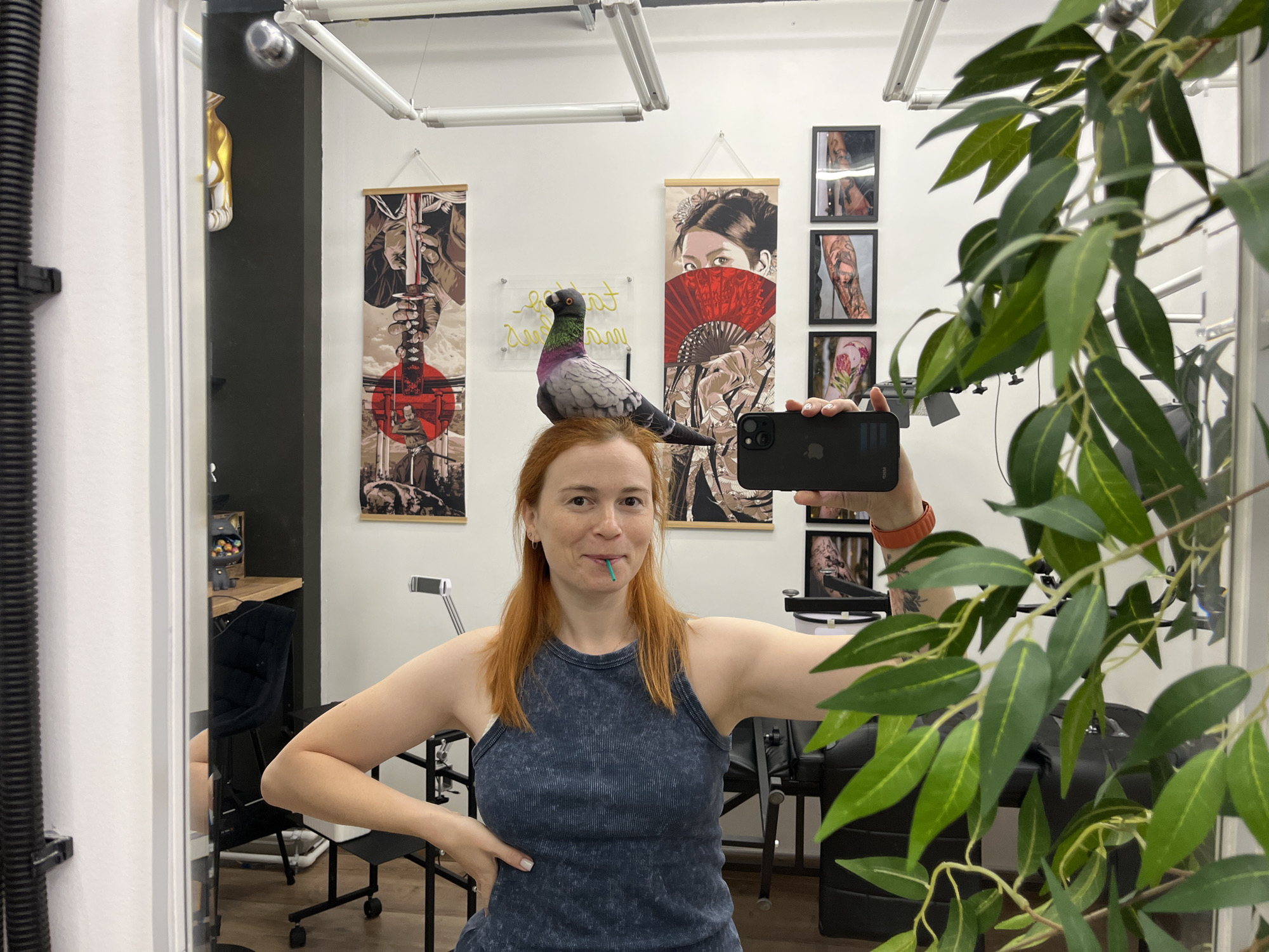 A person with red hair is taking a mirror selfie in a room decorated with Japanese-style art and tattoos. The person is wearing a dark blue tank top and has a green object, possibly a toothpick, in their mouth. A taxidermied pigeon is perched on the person's head. The room appears to be a tattoo parlor, with tattoo chairs and artwork visible in the reflection. The overall scene is casual and playful. There is no indication of the city of Chaikovsky, Russia.