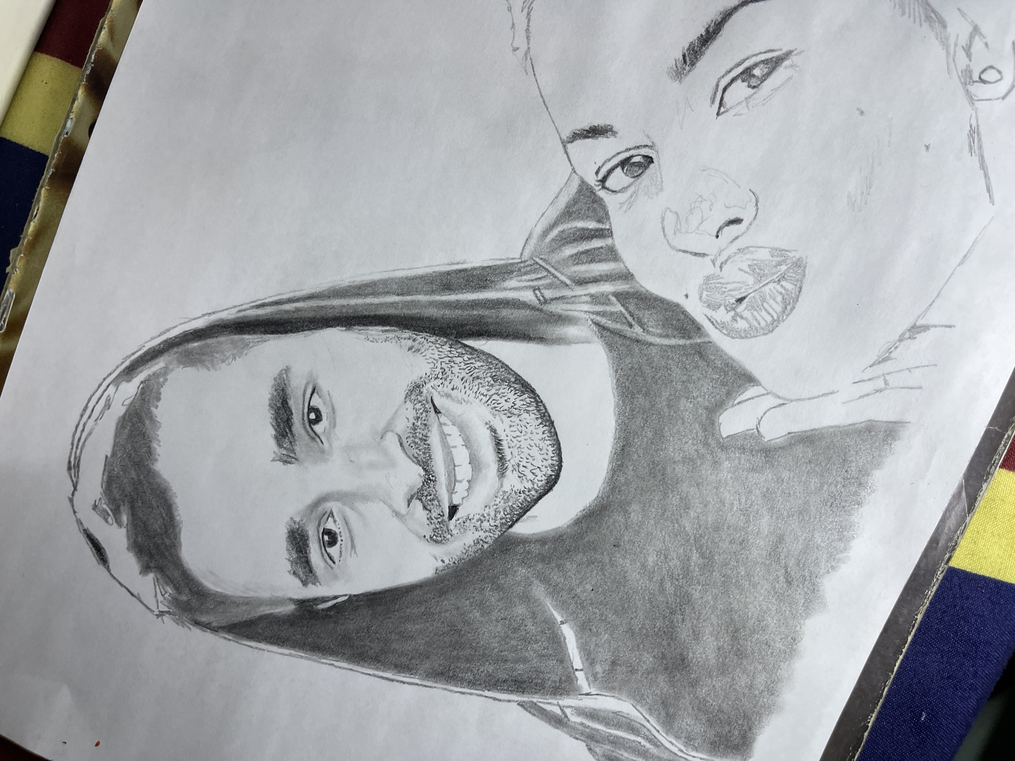 This image displays a black and white pencil sketch on a piece of white paper, photographed in the city of Jos, Nigeria. The sketch features two individuals. The dominant figure, positioned in the upper portion of the frame, is a male depicted front-facing, smiling broadly with visible teeth. He has short facial hair (stubble) and wears a dark-shaded hooded garment with the hood covering his head. His eyes are dark and his eyebrows are prominent. Partially visible in the lower right, the head of a female figure is sketched, tilted at an angle, with her eyes directed towards the viewer. Her lips are full and slightly parted. Her face is rendered with lighter shading compared to the male figure, and a lightly outlined hand is positioned near her chin. The white paper shows minor creases. The bottom edge of the paper rests upon a worn, dark-colored object resembling a book or folder binding, beneath which a multi-striped fabric with red, yellow, and blue bands is visible.