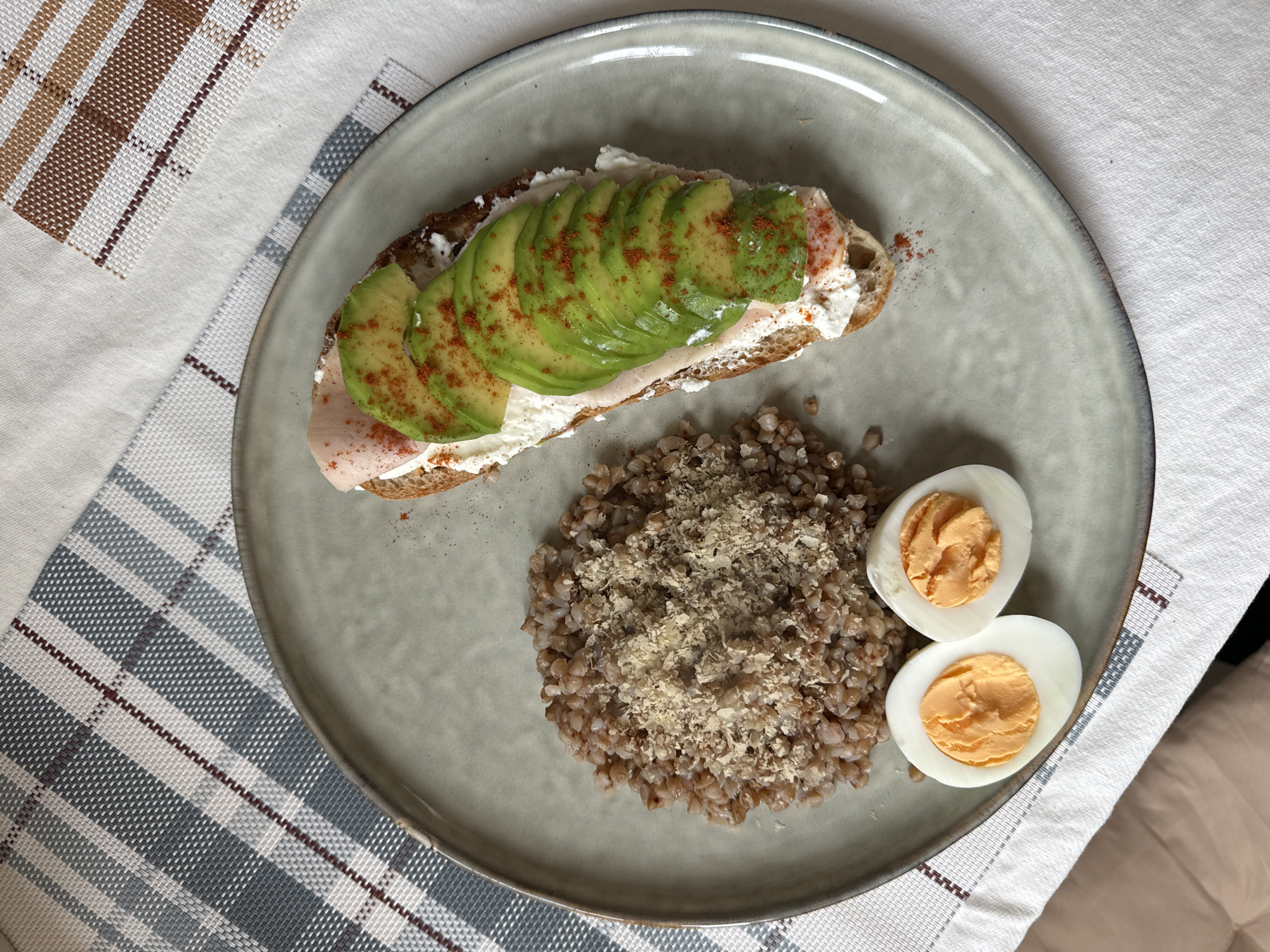 An overhead shot reveals a prepared meal served on a round, light gray ceramic plate with an irregular rim. The plate contains three distinct components. On the upper left, a mound of cooked buckwheat groats is topped with a finely grated, light brown substance. Directly below the buckwheat, two halves of a hard-boiled egg are present, cut lengthwise to expose firm, orange yolks. To the right, an open-faced sandwich occupies a significant portion of the plate. It is constructed on a slice of dark bread, topped with a generous layer of white, creamy spread. Above this spread, a slice of light pink deli meat is visible, covered by multiple thin, fanned slices of green avocado. A reddish powder, consistent with paprika or chili flakes, is sprinkled over the avocado and parts of the creamy spread. The plate rests on a white fabric surface, with a checkered placemat visible beneath and to the left, featuring a pattern of light blue, white, and thin dark red stripes. Another section of a placemat with brown, white, and darker brown checks is partially visible to the upper right. The scene is presented as a domestic meal setting within the city of Krasnoyarsk, Russia.