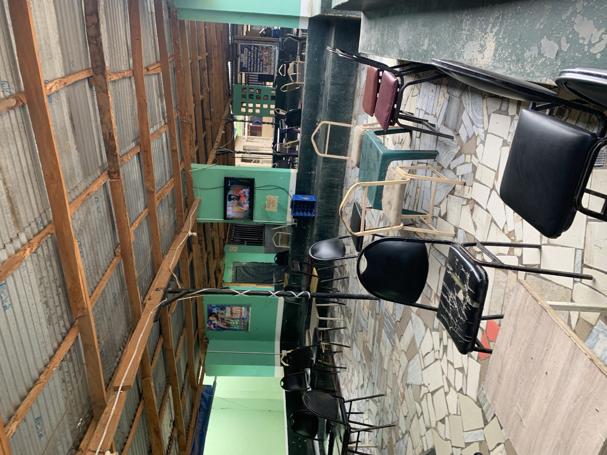 The image depicts an open-air restaurant or eatery in Kabong, Nigeria. The seating area features a mosaic tile floor with a mix of black and metal-framed chairs, some with padded seats. Tables are simple, wooden, and rectangular. The restaurant has a corrugated metal roof supported by wooden beams. A television screen displays a video. A sign advertising "Kitchen, Cat Fish Pepper Soup, Goat Peppered Meat & Soup, Plantain & Scrambled Eggs, Chicken Popper, Grilled Fish" is visible. The structure is painted in shades of green.