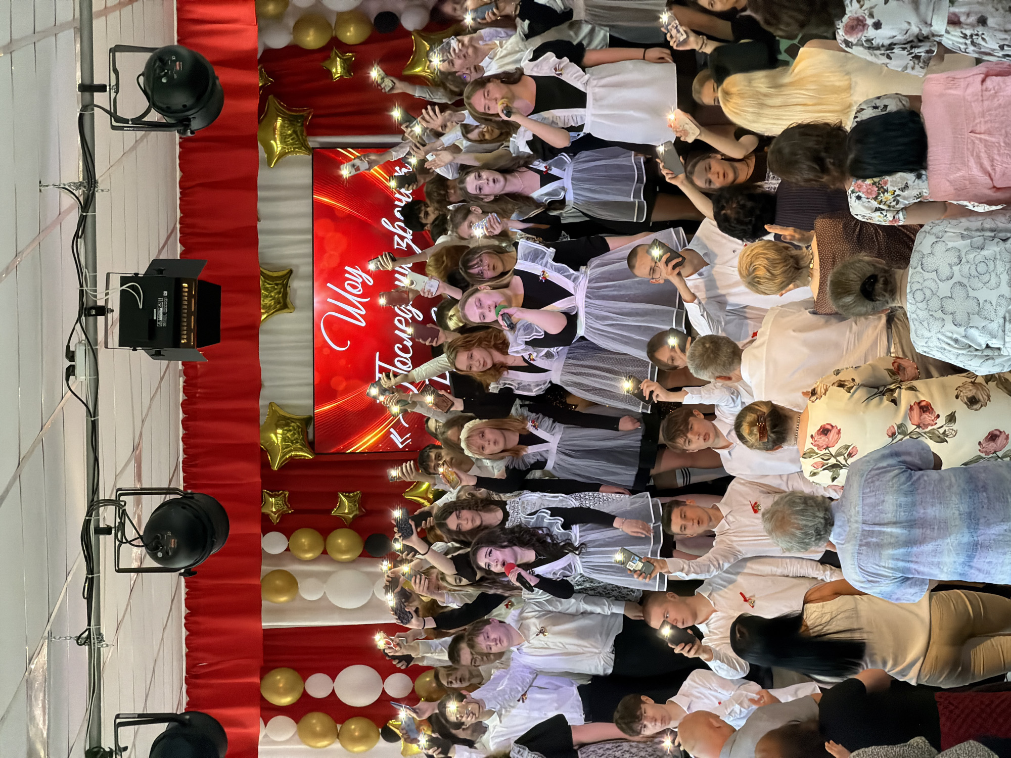 A group of graduating students in Izhevsk, Russia, are onstage with microphones, likely performing. The students wear black and white school uniforms. Many are holding up phones with the flashlights on. A red and white backdrop is visible behind them, decorated with gold and white balloons and star-shaped decorations. Stage lights stand to the side. Some audience members are also visible in the foreground.