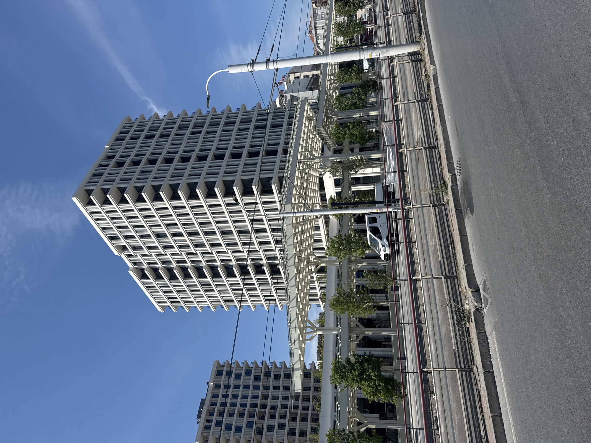 The image captures a modern architectural scene in Bahçelievler, Turkey, under a clear blue sky with wispy clouds. The foreground shows a paved road with a median strip and a metal railing. Beyond the railing, a multi-level structure with white columns and a canopy is visible, suggesting a pedestrian walkway or a commercial area.
Dominating the midground are two tall, contemporary apartment buildings. The building on the right is particularly striking, with its facade featuring numerous vertical white elements that create a rhythmic pattern of recessed balconies and windows. The building on the left is similar in style but appears slightly shorter and further away.
A white van is seen driving on a road behind the canopy structure, suggesting ongoing urban activity. The environment is clean and well-maintained, characteristic of a developed city district. The lighting indicates it is daytime, with shadows cast by the structures and trees. The overall impression is one of modern urban development and infrastructure.
