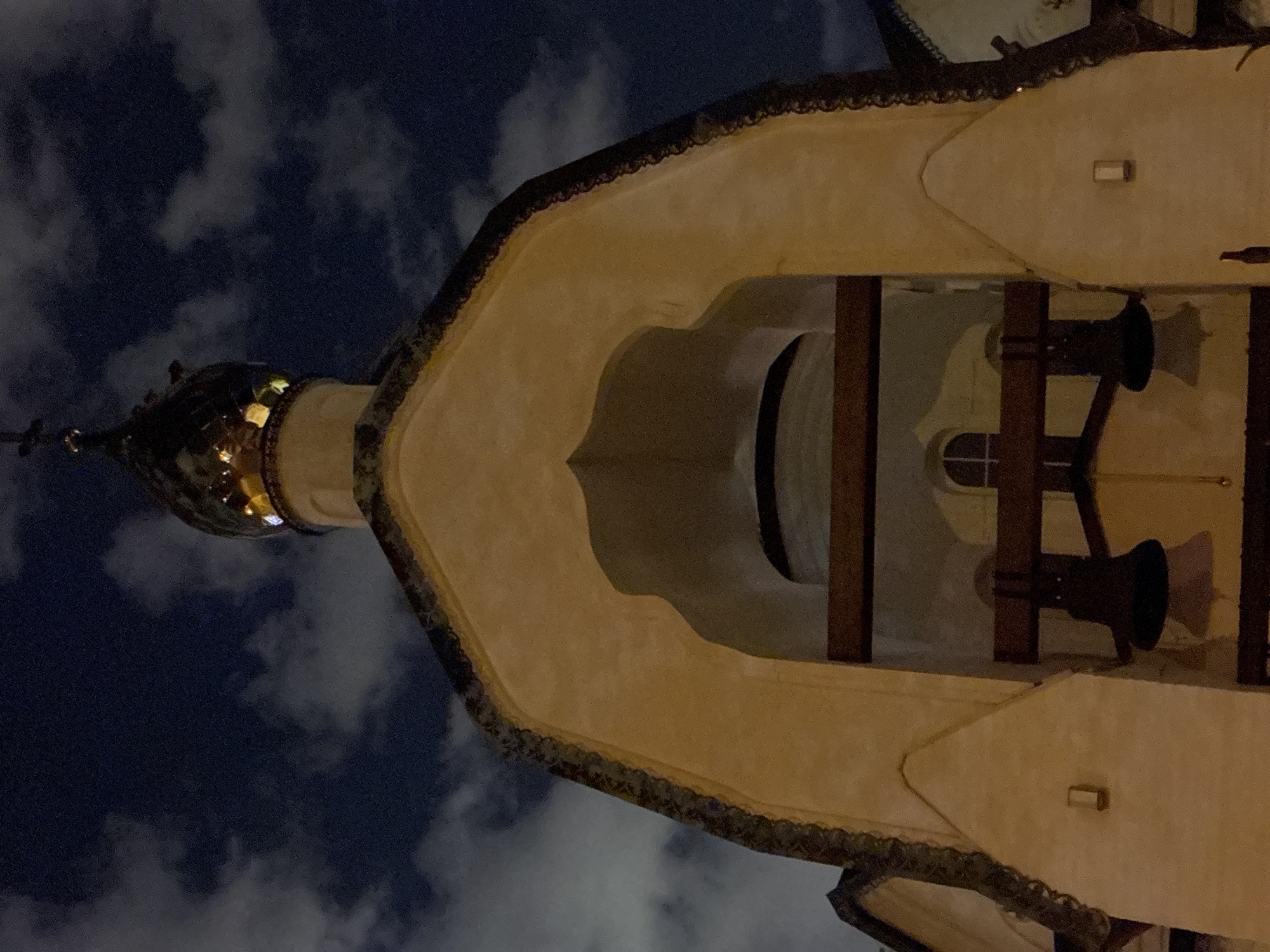 The image shows a low-angle view of the Church of the Intercession at Fili in Moscow, Russia, at night. The building's golden onion dome is visible against a backdrop of dark, cloudy sky. The bell tower's archway, with its beige walls and dark trim, frames the interior space where two dark-colored bells hang suspended by wooden supports. A small, arched window is also visible inside the tower. The scene is dimly lit, likely by artificial light sources.
