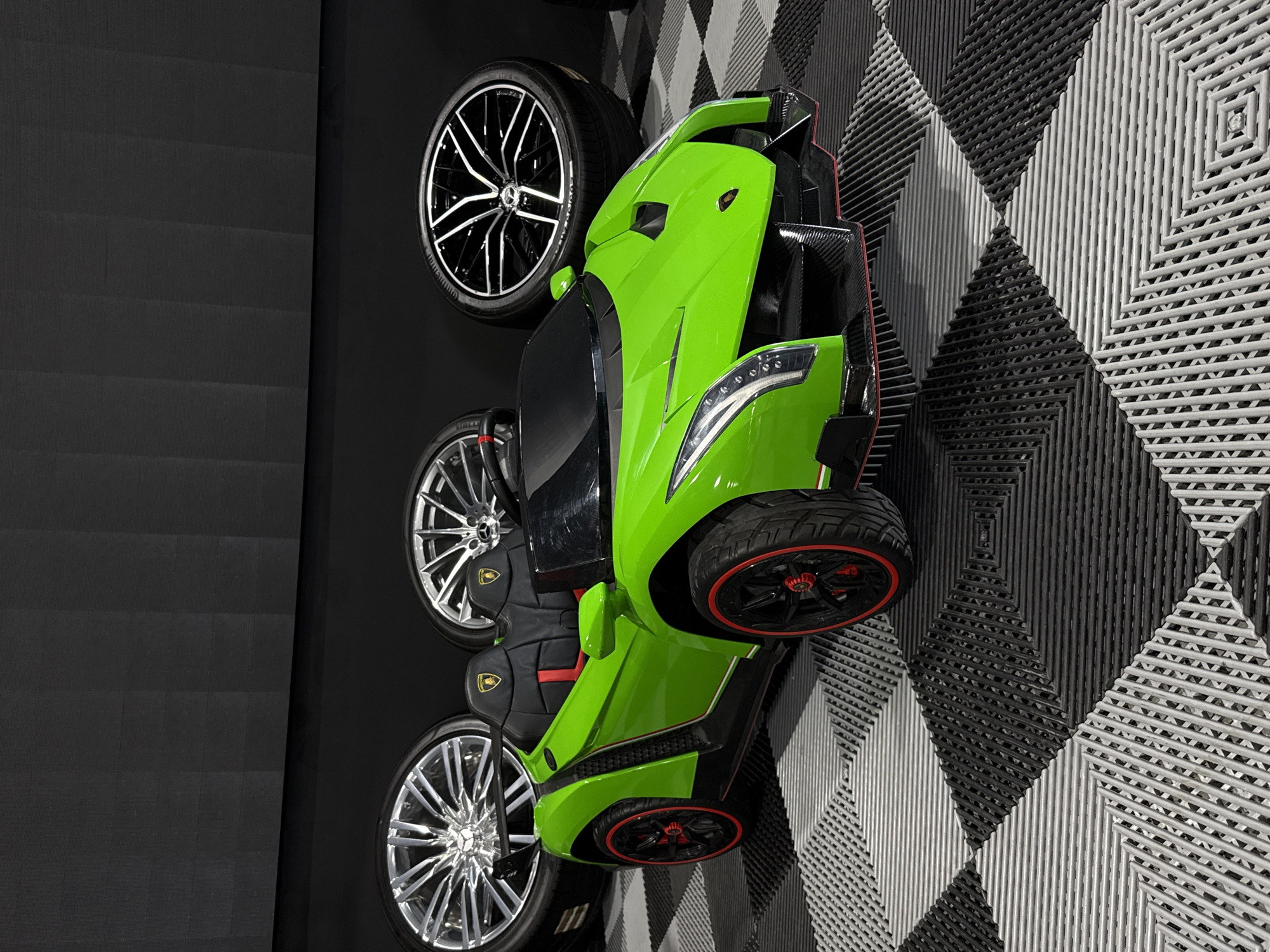 A green Lamborghini toy car is positioned on a wall in Plovdiv, Bulgaria. The car is accompanied by three tires with black rims. The wall is decorated with alternating black and white diamond-shaped tile patterns. An adjacent wall has black tiles.