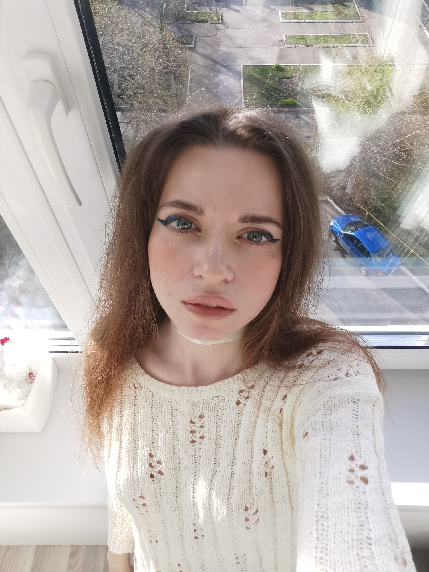 A young woman is taking a selfie by a window. She has long brown hair and is wearing blue eyeliner and a white, textured sweater. Outside the window, a blue car is visible parked on the street. The view from the window includes trees and a building.