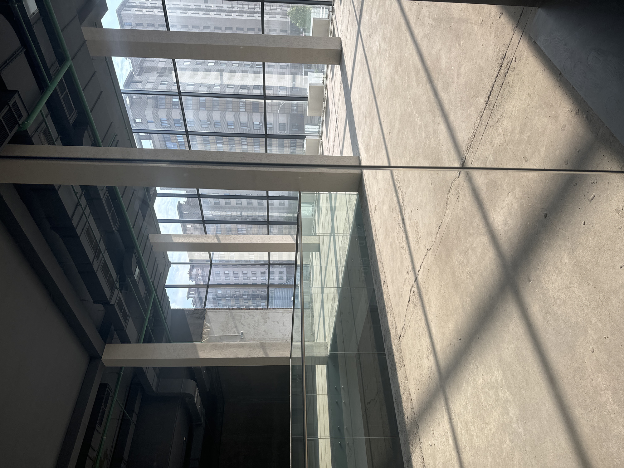 The image captures an interior perspective looking upwards through a grid-like structure of beams and glass windows. The beams appear to be made of concrete or a similar material, and the glass windows offer a view of a cityscape, revealing buildings with a repetitive window pattern. The interior space itself is under construction, with unfinished concrete walls and floors. Pipes run along the ceiling on one side. The location appears to be an urban area, characterized by dense construction. The light filtering through the windows casts shadows on the concrete surfaces.