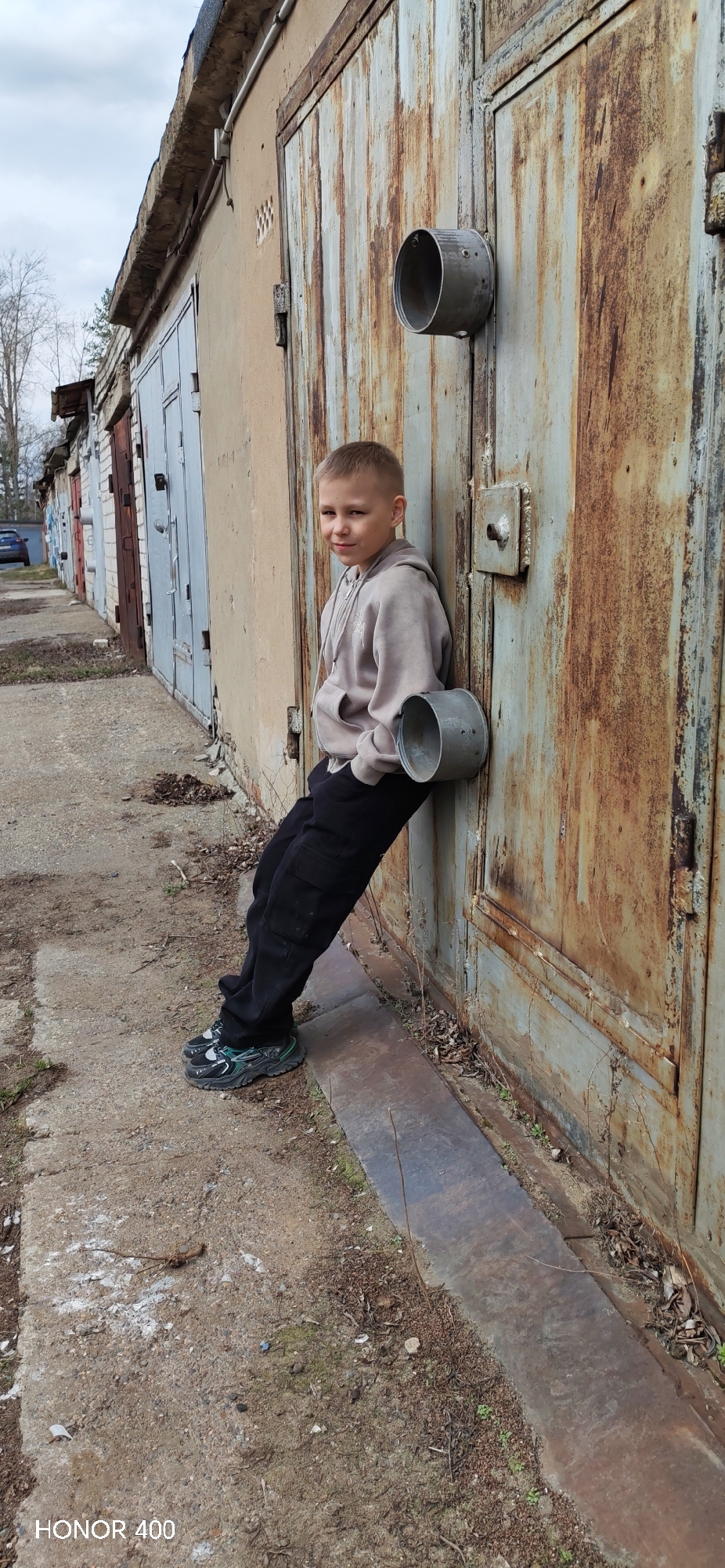 A young boy leans against a rusty garage door at 1/5 Gorkogo Street, Chaikovsky, Perm Krai, Russia, 617763. The boy wears a light tan hoodie and dark trousers, and he has short, light-colored hair. The garage door is part of a row of similar garages, exhibiting varying degrees of weathering and rust. Two cylindrical metal objects are affixed to the garage door, one at chest level and the other above the boy's head. The ground is a mix of concrete, dirt, and debris, with sparse vegetation growing near the base of the garage doors. A line of garages extends into the distance.