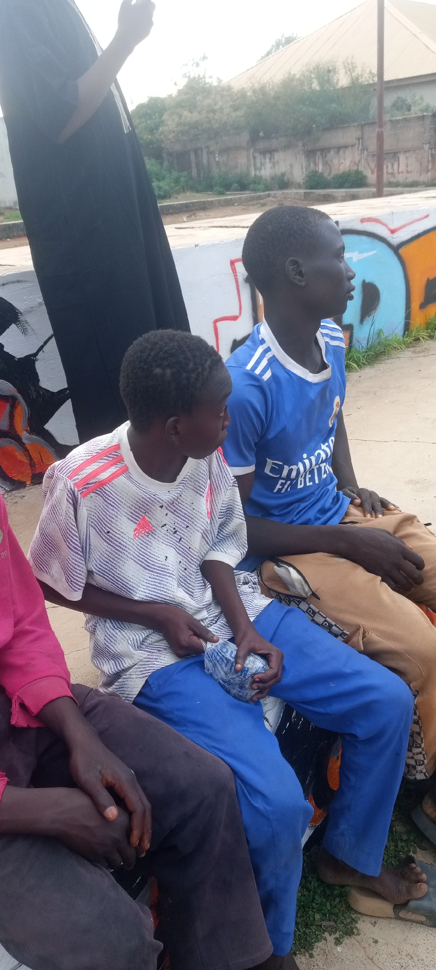 Three youths sit on an elevated concrete structure, possibly a retaining wall or bench, in an outdoor environment. The youth on the left wears a pink shirt and dark pants. The youth in the middle wears a white shirt with pink stripes and blue pants, holding a wrapped object. The youth on the right wears a blue soccer jersey and brown pants. They are all looking to the right. Behind them, there's a wall with graffiti, and beyond that, vegetation and buildings. The location appears to be urban but lacks specific identifying details.