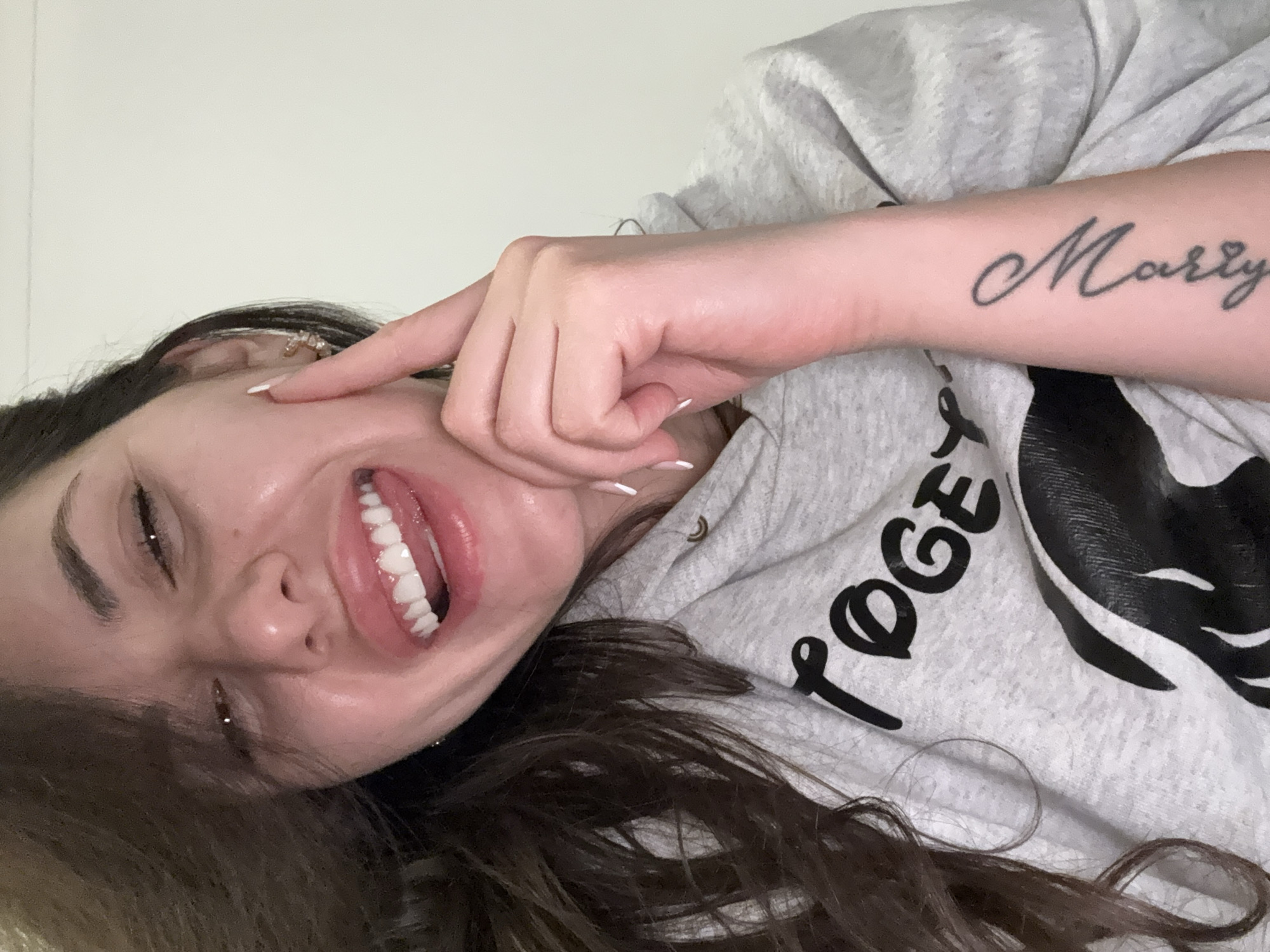 A woman with long, dark hair smiles broadly, her teeth visible. She is wearing a light grey hoodie with black lettering. On her left forearm is a tattoo that reads "Marry." Her right hand is raised, her index finger touching her cheek. The background is a plain white wall. There are no clear indicators in the image that identify this location as London, United Kingdom.