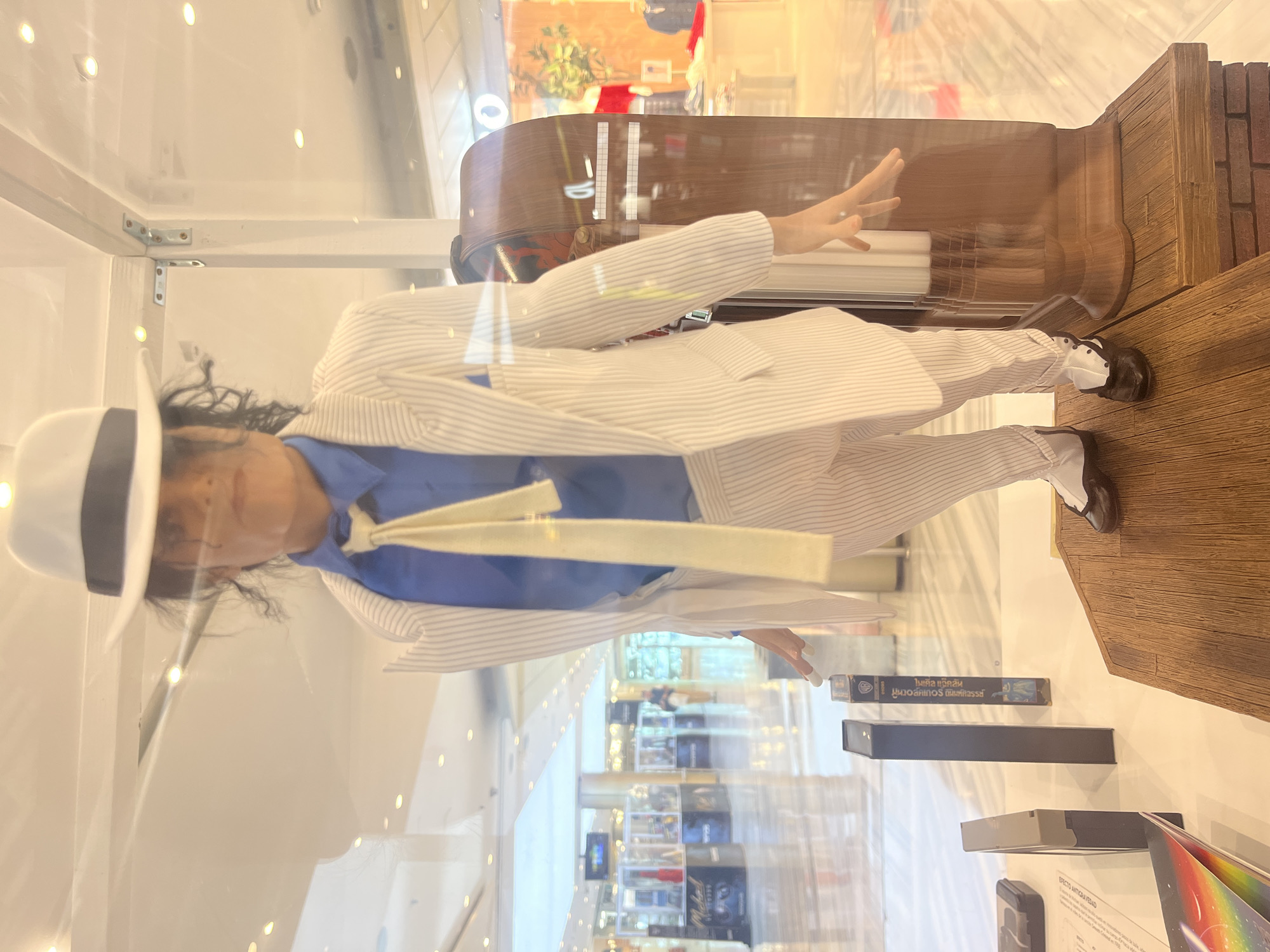 An intricately detailed mannequin of Michael Jackson, dressed in his iconic white pinstripe suit, blue shirt, white fedora, and two-tone shoes from the "Smooth Criminal" era, is displayed upright within a clear glass case. The figure is posed in the famous anti-gravity lean, with one leg bent and the torso tilted sharply forward. Behind the figure, a dark wooden structure resembles part of a grand piano or a stage set. The scene is set indoors, likely within a shopping mall or exhibition space in Las Palmas, Spain, as indicated by the reflective ceiling lights and blurred reflections of other stores or areas visible through the glass.
Inside the display case, below the figure's feet, are various objects, including several dark rectangular items resembling VHS tapes or books. One visible item is a book or magazine featuring a vibrant rainbow graphic.
Visible text on an object inside the case includes Thai script: "ไมเคิล แจ๊คสัน" (Michael Jackson) and "มูนวอล์คเกอร์ ถนิมพ์ศิลรรช" (Moonwalker). Another piece of text in Spanish reads "EFECTO ANTIGRAVEDAD" (Anti-Gravity Effect), directly referencing the figure's pose and Michael Jackson's signature move. The overall presentation is a tribute to the pop icon's legacy.
