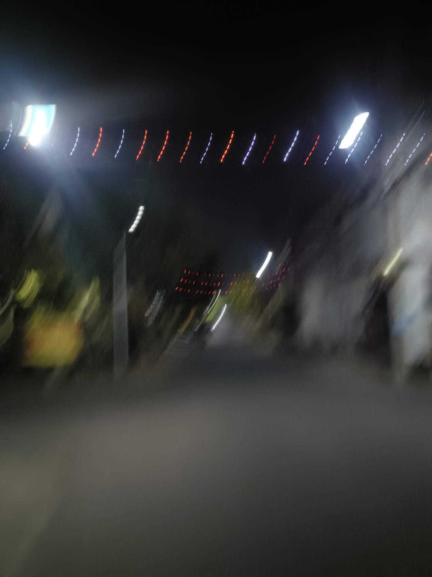This is a motion-blurred, low-light photograph taken at night in Kediri City, Indonesia. The image captures a street scene with blurred lights and shapes, suggesting movement or a slow shutter speed. In the upper portion of the image, there are streaks of light in red, white, and blue, likely from festive decorations or vehicle lights. A bright white light source is also visible on the left side. The foreground consists of a dark, indistinct road surface. In the background, there are blurred shapes of trees and buildings, with further streaks of light suggesting the presence of other streetlights or illuminated structures. The overall impression is one of a busy street at night, possibly during a celebration or event, rendered abstract by the motion blur.