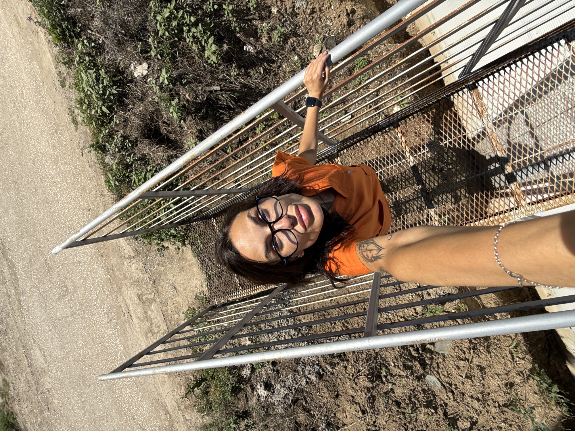 A woman with dark hair and glasses is taking a selfie while standing on a metal staircase outdoors. She is wearing an orange top and has a tattoo on her arm. The staircase appears to be made of rusted metal grates and bars. The background consists of dry, dusty ground with sparse vegetation and a gravel path. The scene is set in an outdoor, possibly urban, environment. The location is identified as Pleven, Bulgaria.