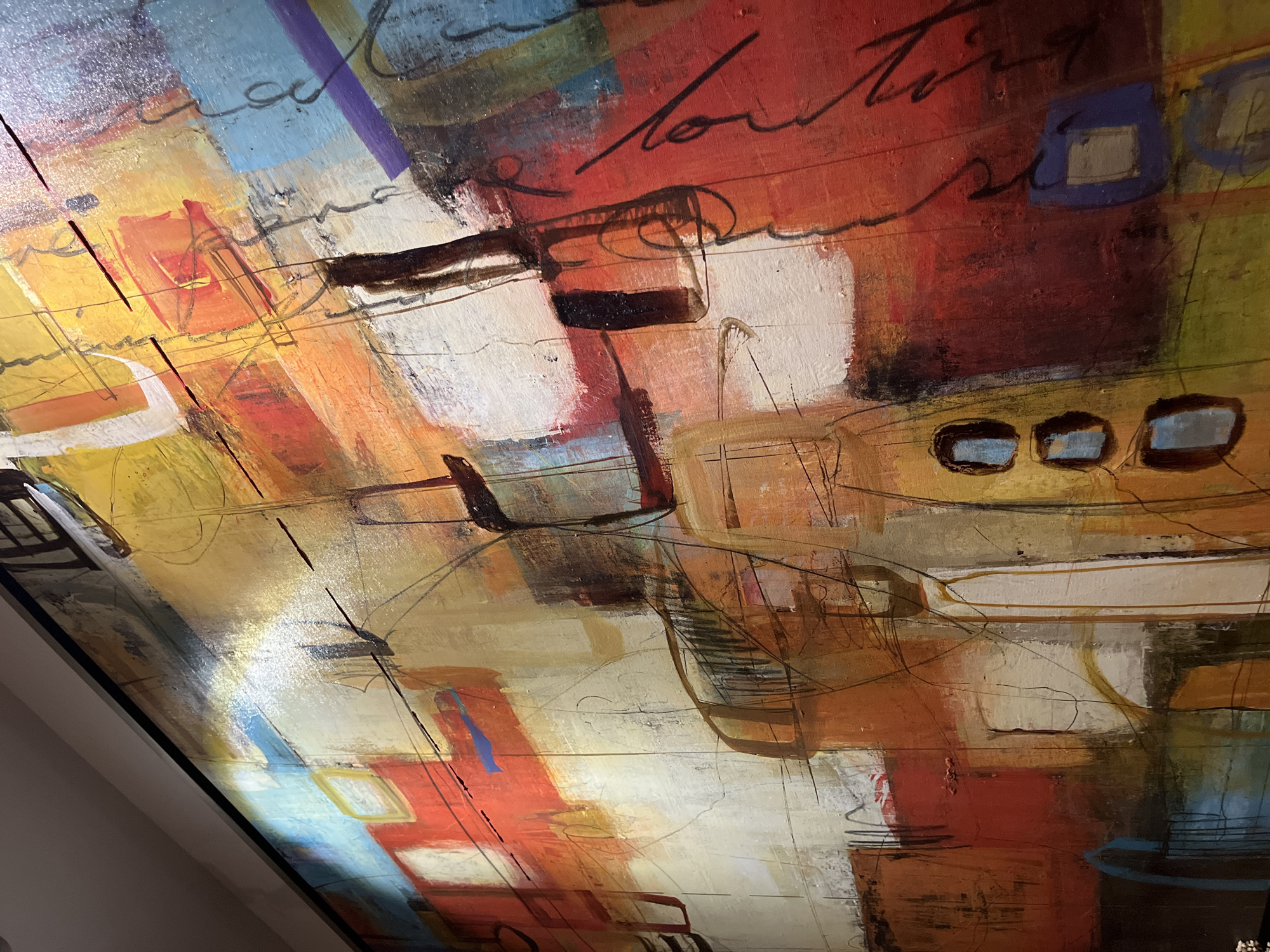 The image shows a close-up section of an abstract painting, likely hanging on a wall. The dominant colors are reds, oranges, yellows, and whites, with accents of blue, green, and purple. The style is characterized by bold, overlapping brushstrokes and linear elements resembling sketched outlines. There's a suggestion of architectural forms, possibly representing buildings or structures, but the interpretation is open due to the abstract nature. Some areas appear to contain calligraphic-like strokes. The painting is mounted within a dark frame, noticeable in the lower left corner.
The location, San Bernardino Tlaxcalancingo, Mexico, is extraneous to the image content as it only depicts a painting. There is no visible evidence to directly tie the image to that location.