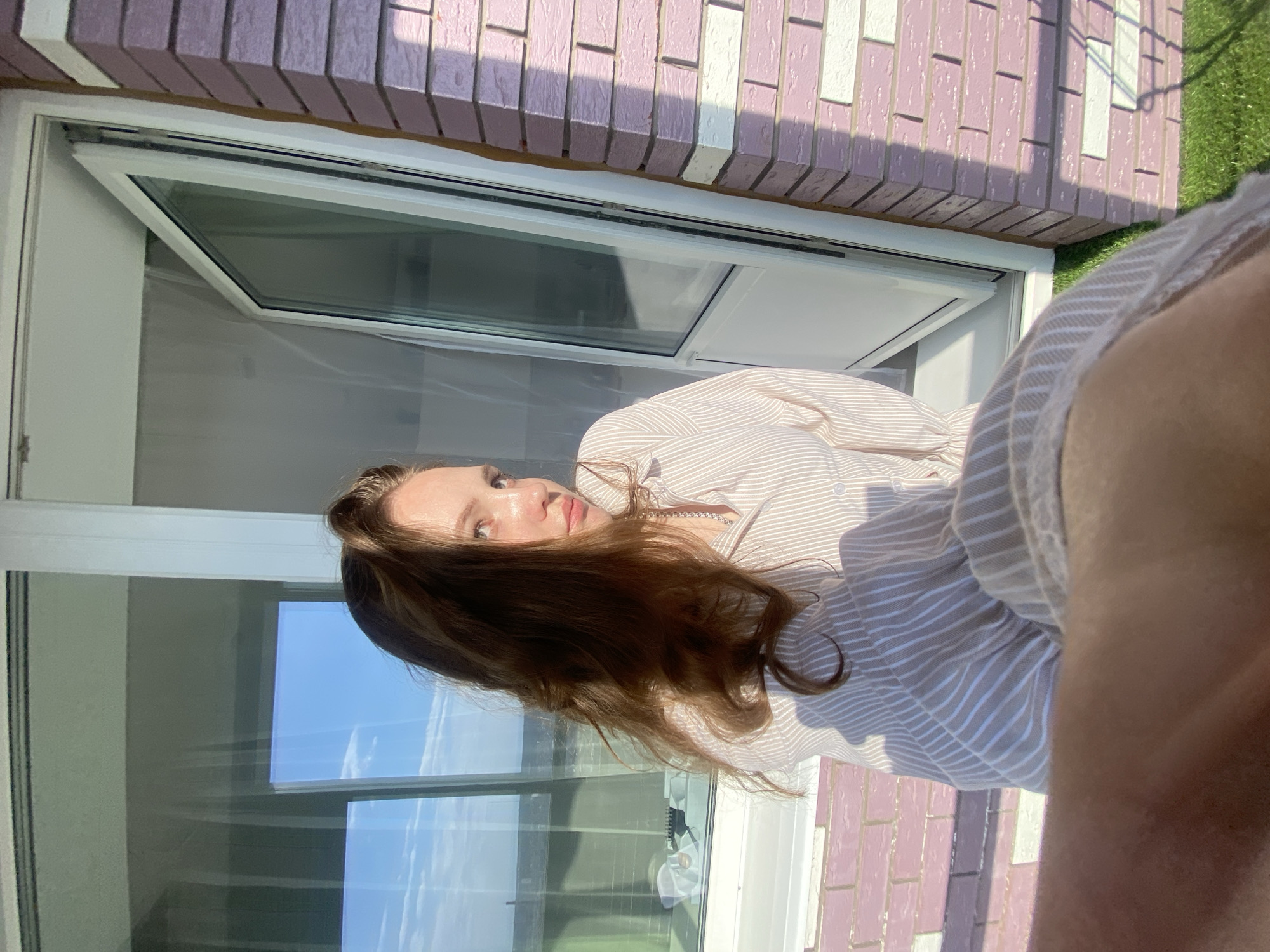 A woman with long brown hair is seen in a vertically-oriented eye-level shot. She is wearing a striped shirt. She is positioned in front of a window. In the reflection of the window, greenery and the sky are visible. The wall outside the window is constructed of pink and white bricks. The scene is possibly located in Izhevsk, Russia.