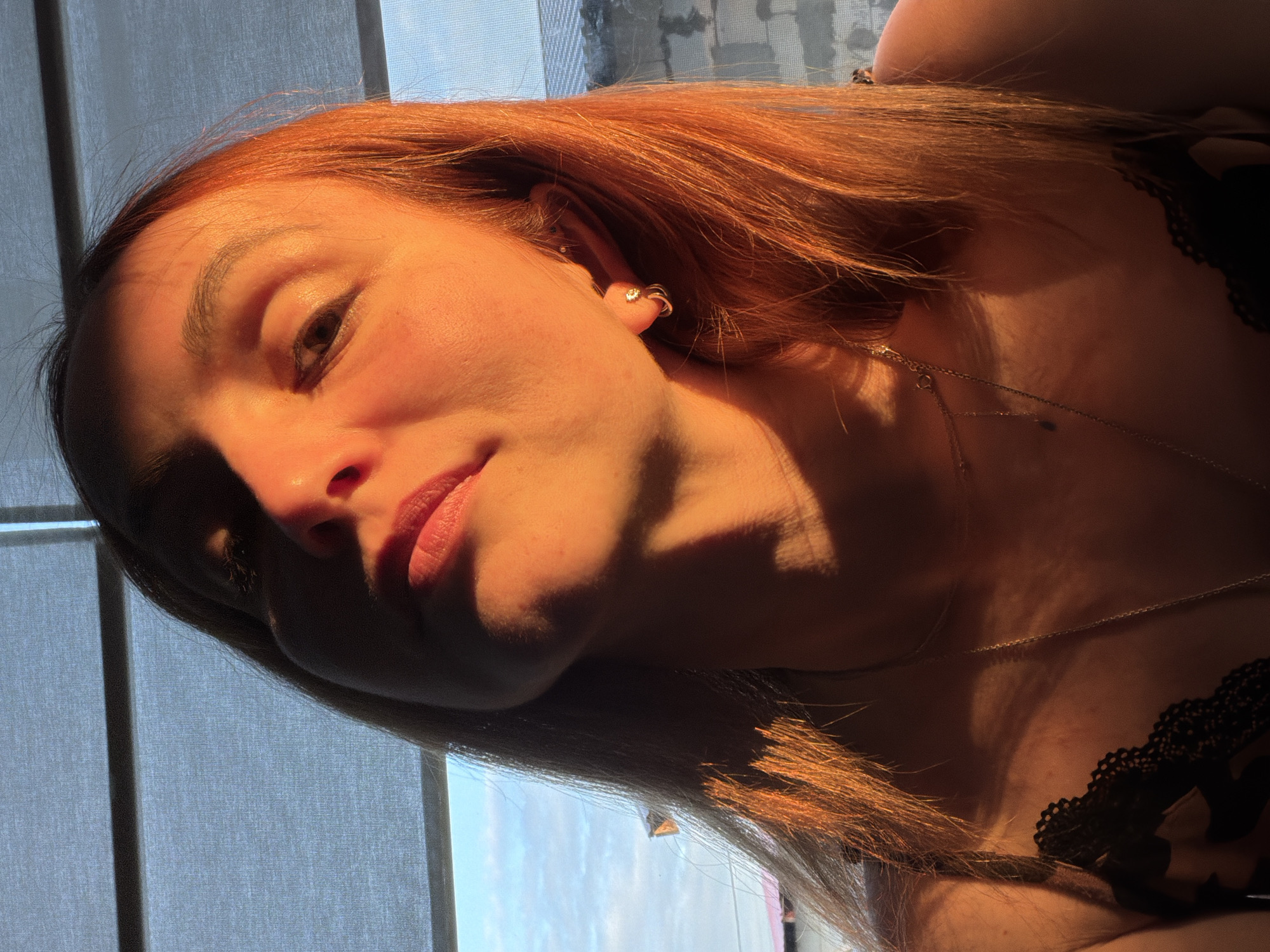 A close-up portrait shows a female subject with reddish-brown hair, her head tilted upwards, illuminated by strong, warm, directional light. The light source, positioned to the subject's upper right, casts prominent shadows across her left facial features, neck, and upper torso, while brightly illuminating her right cheek, nose, and lips. Her right eye displays a catchlight. She is wearing multiple thin chain necklaces and two earrings in her right ear: a hoop with a clear stone and a small stud. Her visible garment has a black lace trim. The background features light-colored window blinds across the top, and through the window on the right, a segment of a blue sky with clouds and a distant building structure, indicative of an outdoor environment in the city of Chaikovsky, Russia.