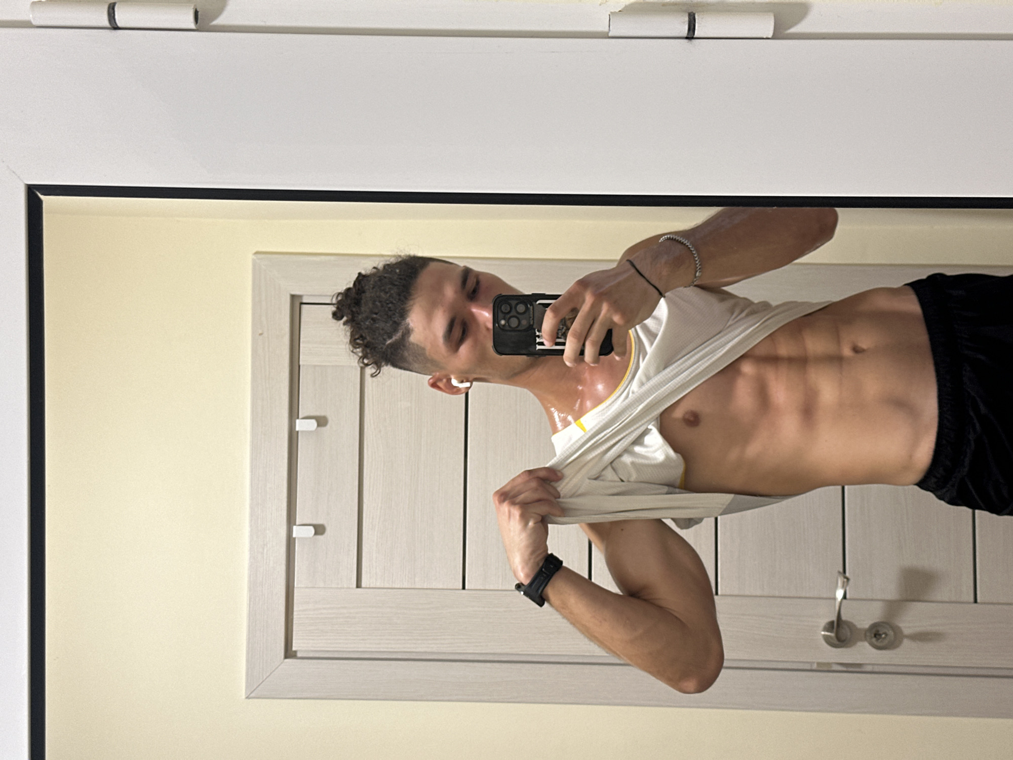 The image is a selfie taken by a young man in what appears to be a room with a light yellow wall and a large closet or cabinet with white wooden doors. The man is wearing black shorts and a white tank top, which he is lifting up to reveal his abs. He has dark, curly hair tied up in a bun and is wearing a silver bracelet on his left wrist and a black watch. He is holding a smartphone in his right hand, and the phone's screen is visible, showing a camera app. The lighting suggests it is daytime, and the man appears to be in good spirits, perhaps showing off his physique. The closet doors have silver handles and what looks like a keyhole. The overall setting is casual and personal.