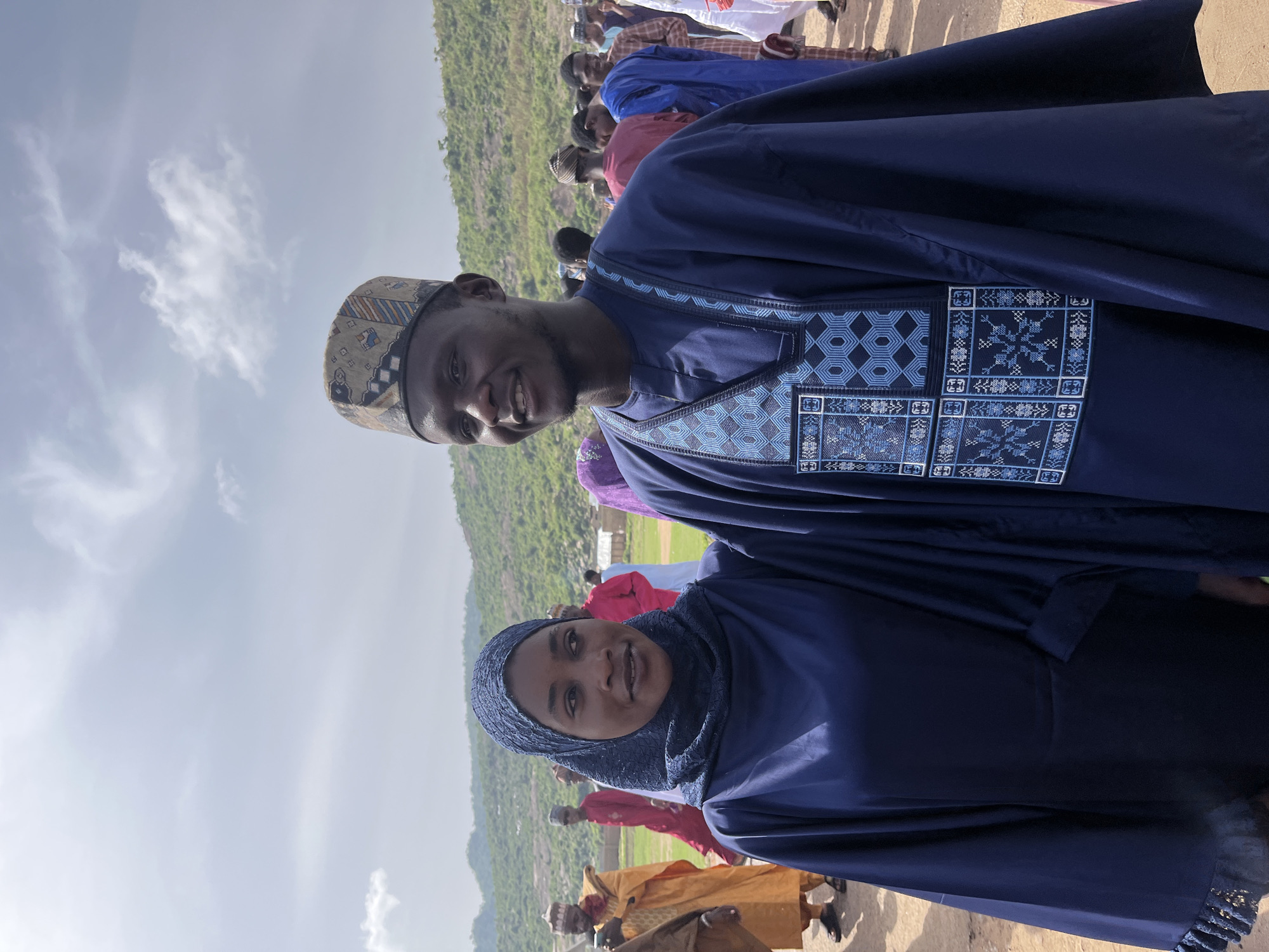 The image captures a man and a woman standing close to each other, both smiling and looking towards the camera. The man is wearing a dark blue traditional garment with intricate embroidery on the chest area, and a patterned cap. The woman is wearing a dark blue hijab and a matching garment. Other people are visible in the background, some dressed in brightly colored clothing. The backdrop consists of a green hillside under a partly cloudy sky. The setting appears to be outdoors in Naraguta Hausawa, Nigeria.