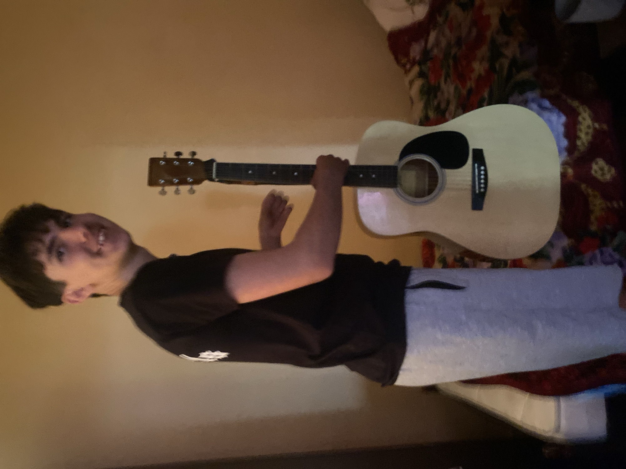 A person is holding an acoustic guitar indoors, sideways. The person has short dark hair and is wearing a dark t-shirt and grey sweatpants. The guitar is light-colored with a dark pickguard. The background includes a wall and floral bedding. There is no indication or evidence of the location being Ustyvytsia, Ukraine.