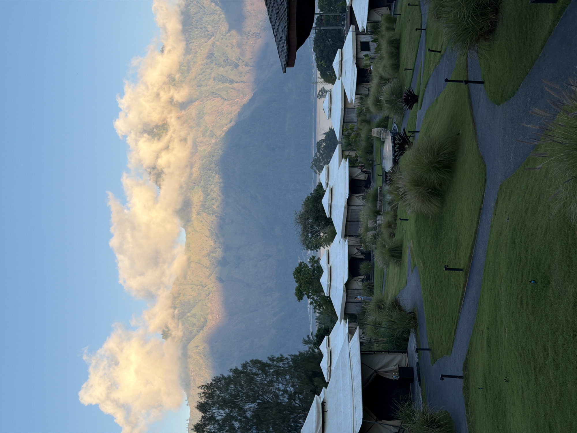 This image depicts a glamping resort in Kintamani, Indonesia, with a backdrop of a majestic mountain range partially obscured by illuminated clouds during either sunrise or sunset. The foreground features several well-maintained canvas tents with white conical roofs, arranged in a neat row on a lush green lawn. Winding asphalt pathways with small, dark lampposts guide visitors through the grounds, which are landscaped with ornamental grasses and low-lying plants. In the middle ground, a circular water feature is visible, surrounded by more vegetation. Further in the background, trees and a body of water are seen before the imposing mountains rise, their slopes textured with shadow and light. A few people are discernible near the tents, one standing and another potentially seated. The sky is a clear blue with streaks of warm light reflecting off the clouds.
