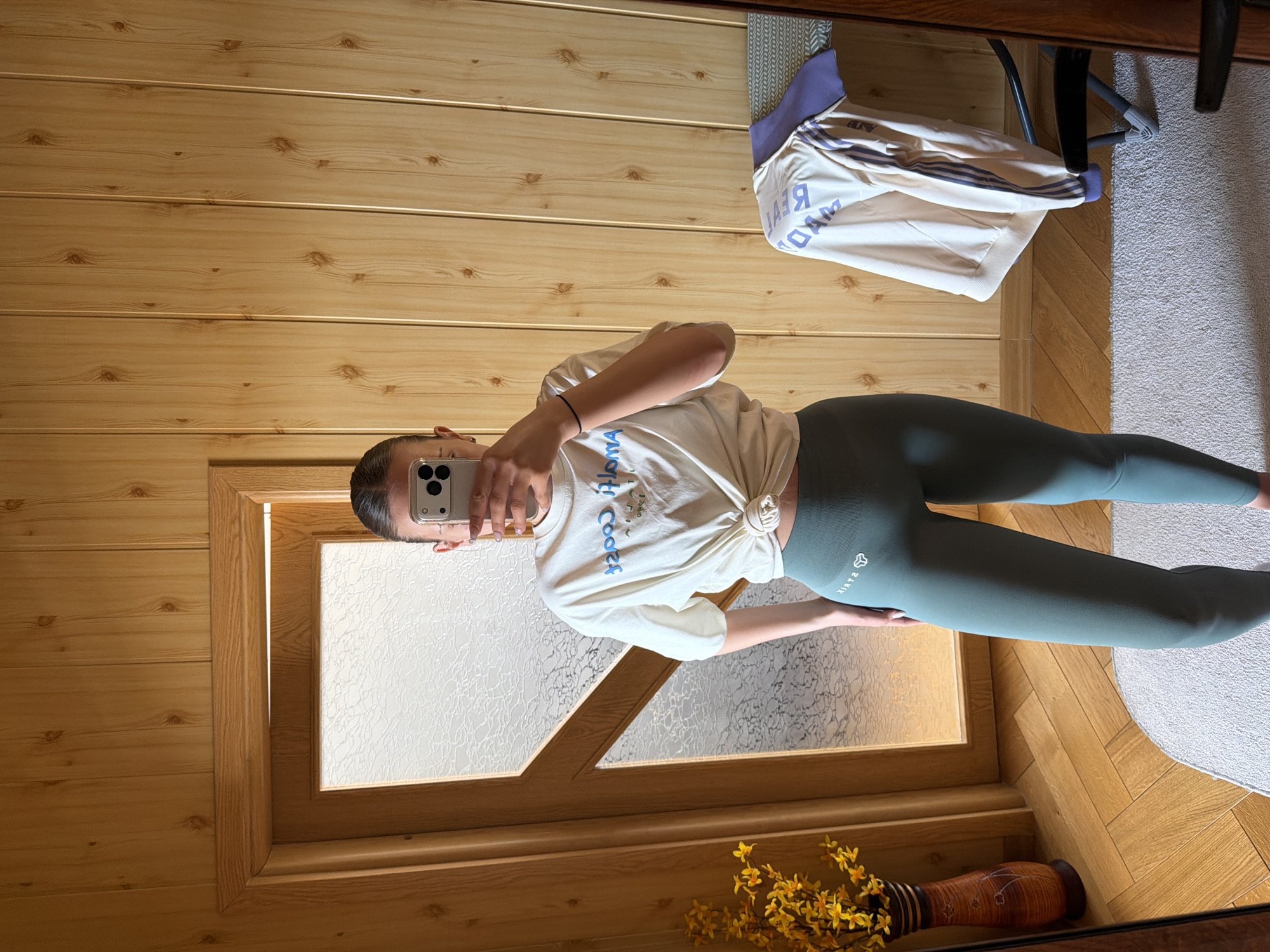 A person is taking a mirror selfie in what appears to be an indoor setting, likely a home in Buhovo, Bulgaria. The individual, seen from the waist up, wears a light-colored, off-white short-sleeved t-shirt tied in a knot at the waist, and dark green or teal full-length leggings with a visible logo on the left thigh. Their hair is pulled back, and they are holding a smartphone with a multi-lens camera system to capture the image, partially obscuring their face. A black elastic band is visible on their left wrist.
The background features light-colored wooden paneled walls and a wooden parquet floor. To the left, a wooden-framed structure, possibly a window or an interior door with frosted or patterned glass, is visible. On the right side of the frame, a section of a light-colored, possibly grey or off-white, textured rug or carpet is present. In the upper right corner, a white sweatshirt or hoodie with purple stripes on the sleeves and purple trim is draped over a dark-framed chair. A tall, patterned brown vase containing yellow flowers is placed on the floor in the lower right corner.
Visible text on the person's t-shirt, appearing reversed in the mirror, shows parts of words like "Coos". The leggings also display a reversed brand logo that reads "STRIK". The sweatshirt in the background has reversed text visible, including "READ" and "MAD". The lighting suggests it is daytime, with ambient indoor light illuminating the scene.