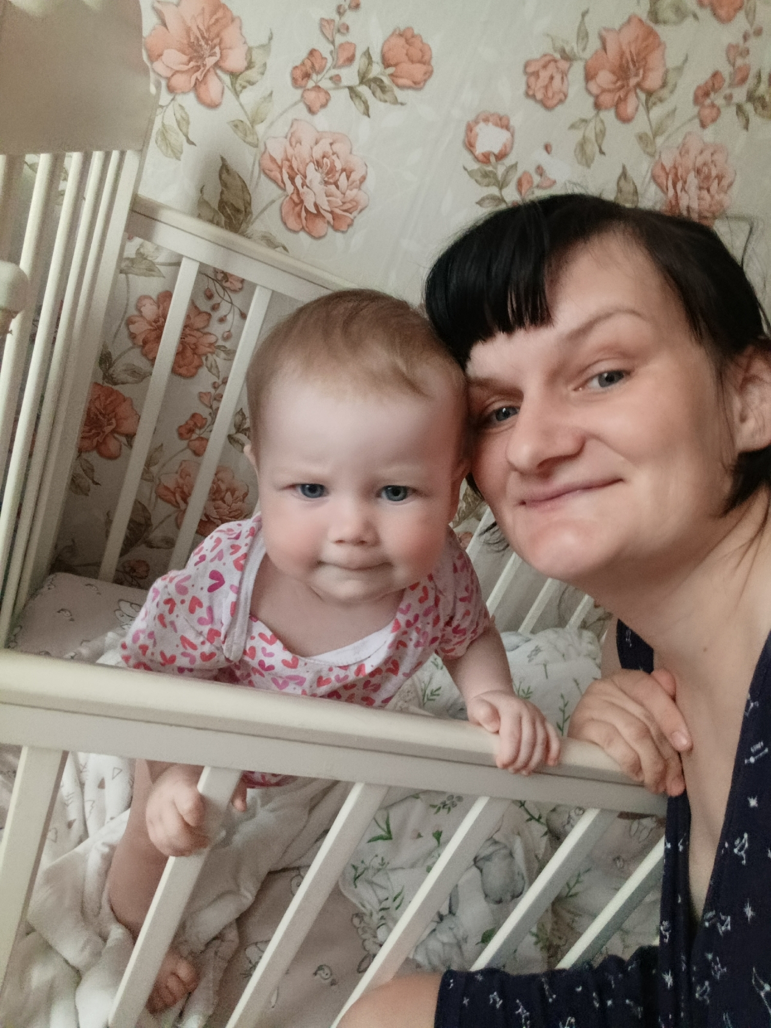 A woman and a baby are in a crib. The woman is on the right side of the image, and the baby is on the left side of the image, leaning on the side of the crib. The crib is white with vertical bars. The baby is wearing a pink and white shirt with hearts on it. The bedding in the crib is white with a print. The wallpaper behind the crib has a floral pattern. The woman is wearing a dark blue dress with white stars. The woman has dark hair with bangs. The location appears to be indoors in a residential setting, but the city and specific location are unknown.