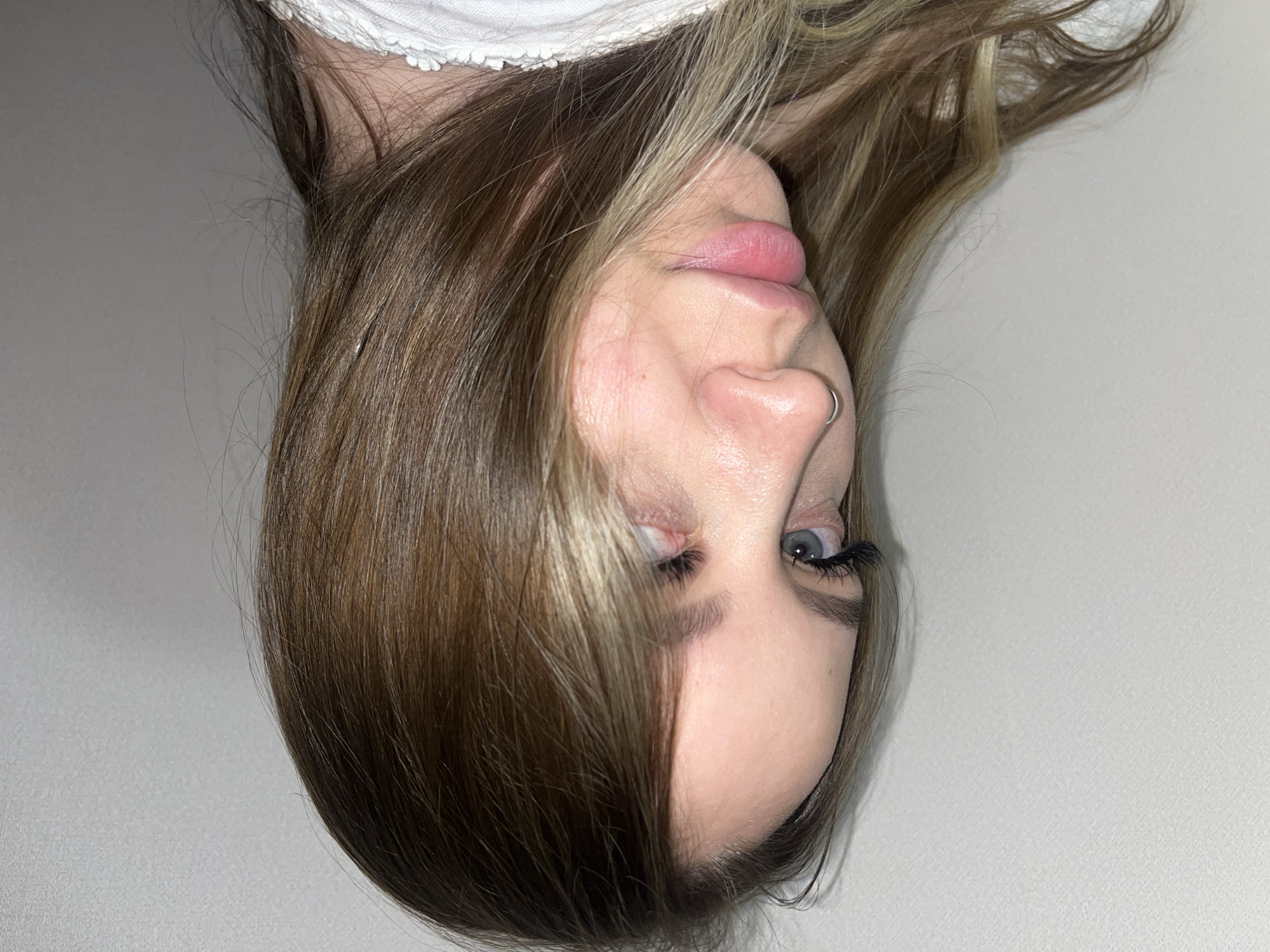 An upside-down medium shot shows the face of a young woman with light skin, brown and blonde hair, and blue eyes. She is wearing false eyelashes and has a silver ring in her nose. Part of a white garment is visible at the top of the frame. The background is a plain white surface. The location is not identifiable as Krasnodar, Russia, from the visual information provided.