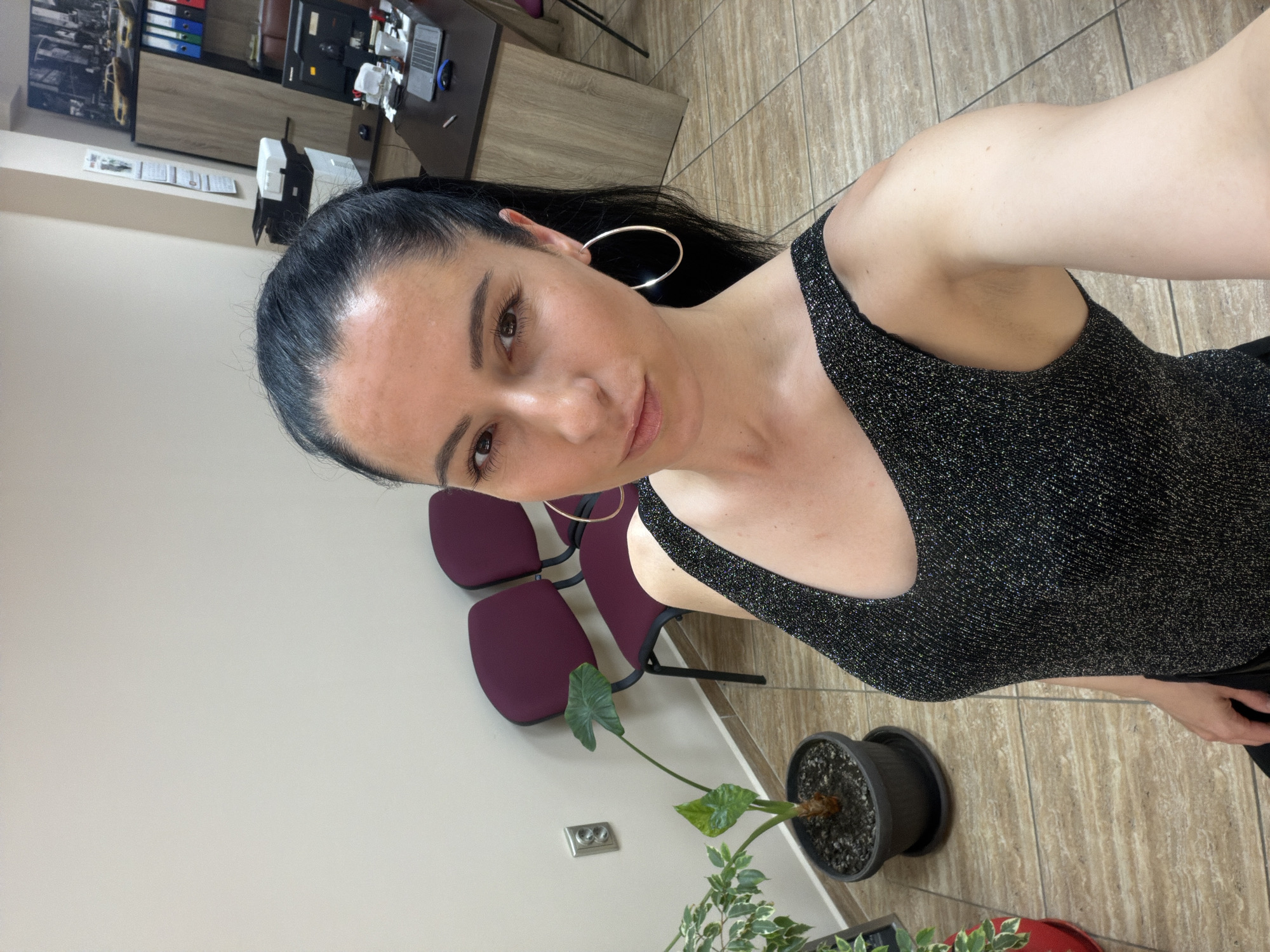 The image shows a woman with dark hair and hoop earrings, wearing a dark, glittery top. She appears to be indoors, in what looks like an office waiting area. In the background, there are purple chairs and a desk with office equipment. A potted plant with large leaves is near the bottom of the frame. Based on the mention of Pleven, Bulgaria, this scene is potentially located in an office within that city.