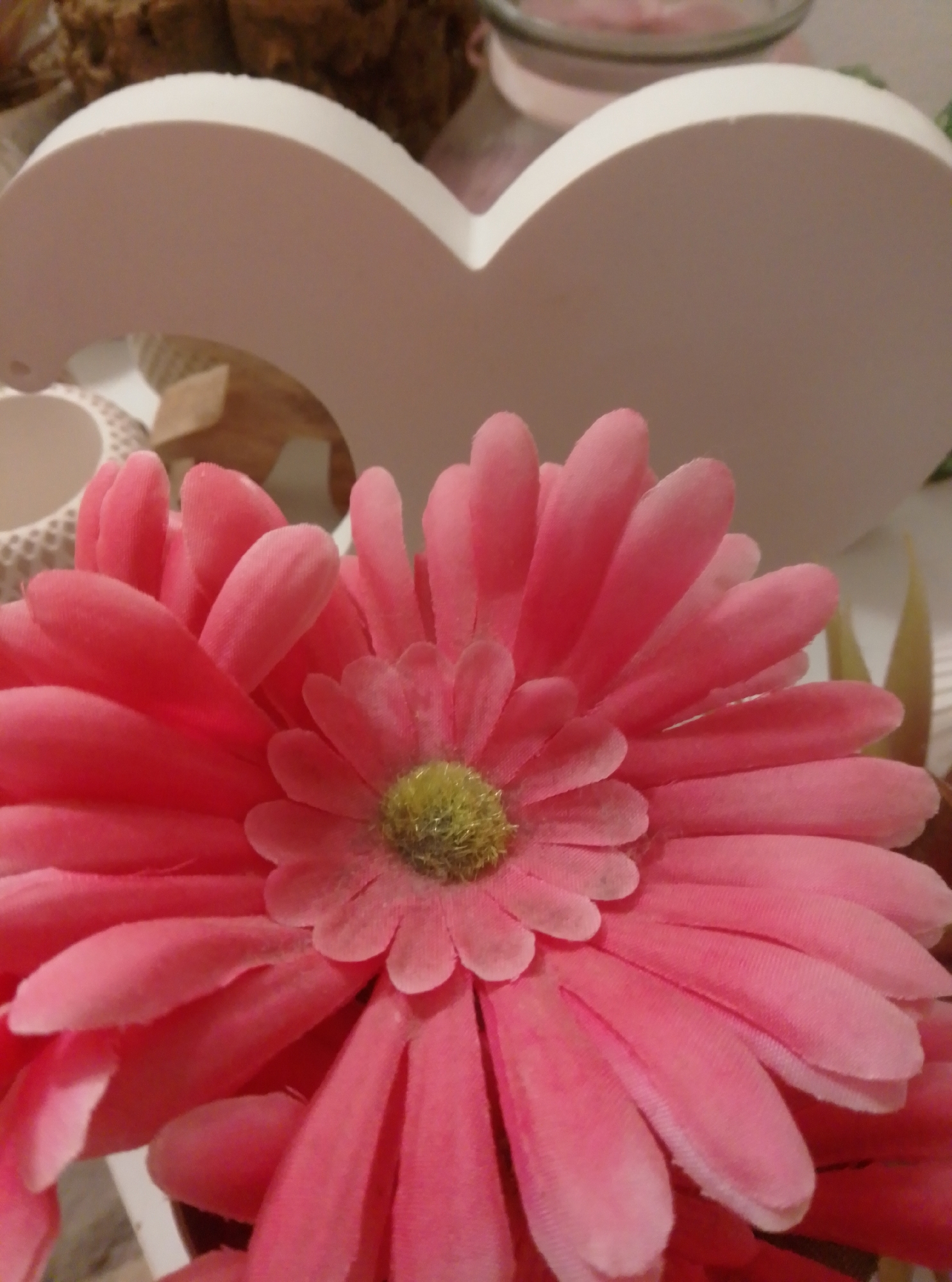 The image shows a close-up of a large, artificial pink gerbera daisy with a fuzzy yellow center. The flower is the dominant subject, filling most of the frame. Behind the flower, a white, heart-shaped decorative object is visible. To the left of the flower, a white patterned container is partially visible, and further back, blurry objects suggest a decorative setting. The background is out of focus, with muted colors and textures. The image appears to be an indoor still life or decorative arrangement. The location in Wedel, Germany, is not discernible from the visual content of the image.