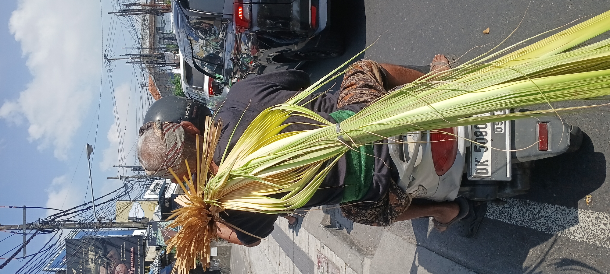 A rear, eye-level, medium shot captures a man riding a scooter in Denpasar, Indonesia, carrying a large bundle of dried palm fronds. The man, with graying hair and wearing a traditional patterned cloth around his head and a green sash over a black t-shirt, is seated on a light-colored scooter. He is transporting the palm fronds, which are tied together and extend significantly behind him, over his right shoulder. The fronds are golden brown at the base and transition to a pale green towards the tips. The scooter’s license plate reads "DK 5080." The scooter is in the right lane of a street, with a paved sidewalk to its left and other vehicles, including a black SUV, a white car, and another vehicle partially obscured, in the background, indicating traffic. Overhead, a dense network of electrical wires is visible against a bright blue sky with scattered white clouds. Commercial signage, including one with the word "Nytman," is present on buildings lining the street. The environment appears to be an urban street scene in Denpasar.