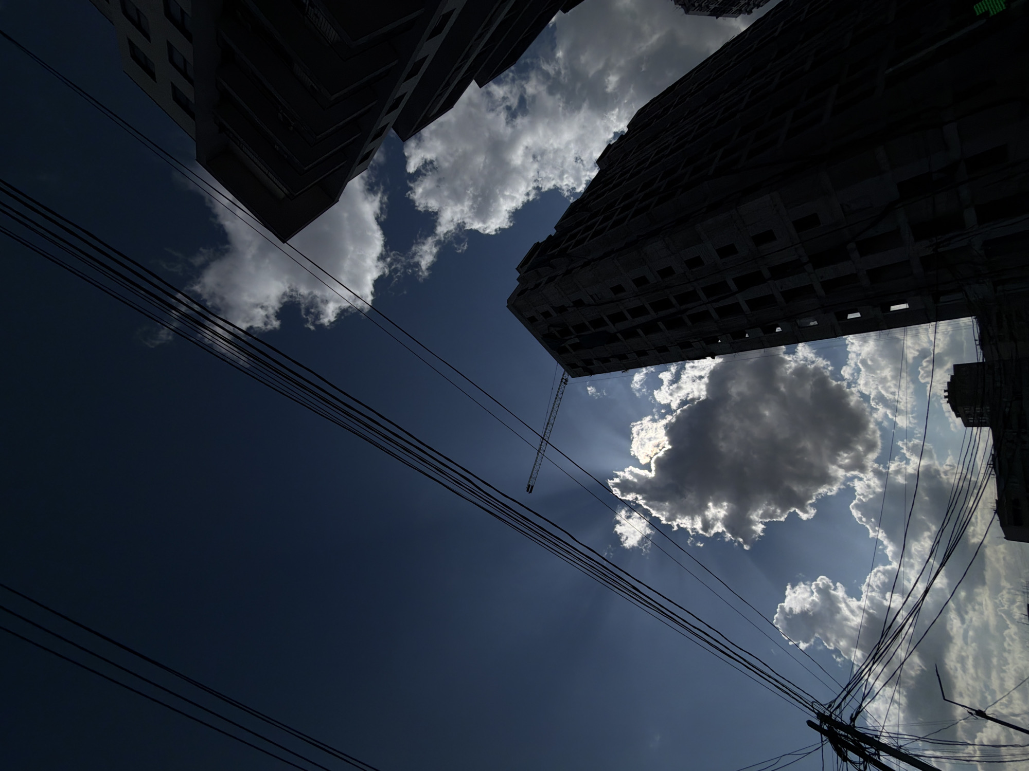 The image presents a low-angle view of the sky in Yerevan, Armenia, framed by tall buildings. The buildings are silhouetted against the bright sky, which is partially obscured by clouds. Power lines crisscross the scene. A construction crane is visible in the distance.