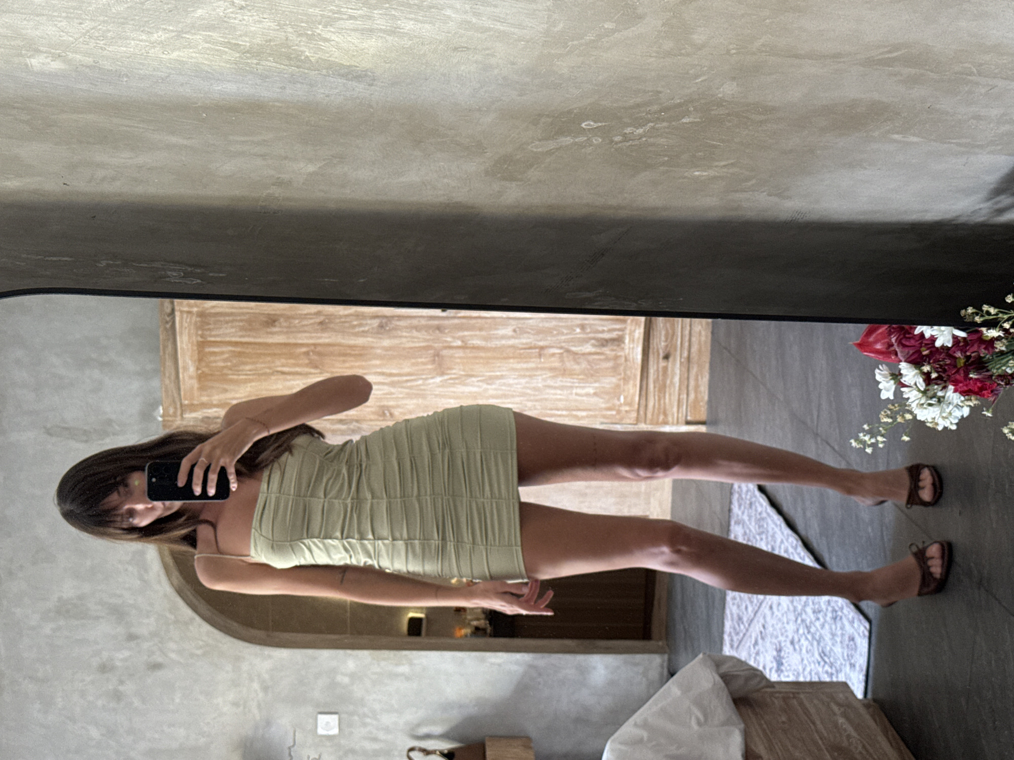 A woman wearing a green mini dress takes a selfie in a mirror in a building in North Kuta, Indonesia. The mirror's frame is dark gray and reflects a light wooden door and gray walls. In the bottom right corner, a vase of red and white flowers sits on a dark gray floor, next to a patterned white and gray rug. A white sheet is partially visible near a wooden table. The woman has long brown hair and is holding a black smartphone. She is wearing brown sandals. A light switch is visible on the wall behind her.