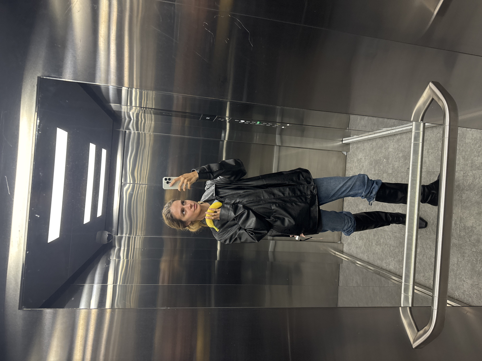 A woman in a black jacket, blue jeans, and black boots holds a yellow banana and a white smartphone while standing in a stainless steel elevator. The elevator has a handrail, floor indicator buttons, and overhead lighting. The location is Unknown, Unknown.