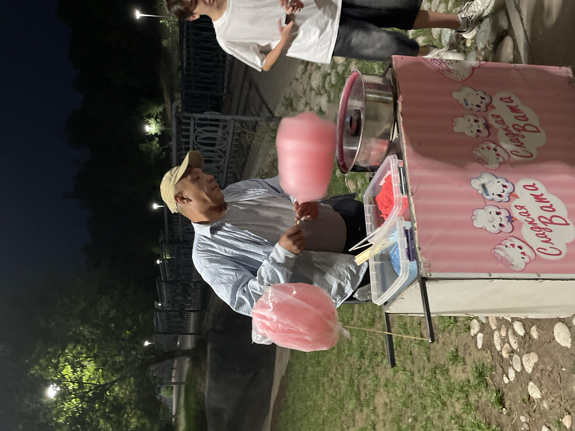 A middle-aged man, wearing a light blue collared shirt and a beige baseball cap, is making cotton candy at an outdoor stand. He is in the process of spinning a large pink ball of cotton candy onto a stick. Next to him, a teenage boy in a white t-shirt and dark pants stands observing. The cotton candy stand is pink and white with cartoon cloud graphics and the Russian words "Сладкая Вата" (Sweet Cotton) printed on it. Several containers of colored sugar, likely for cotton candy, are visible on the stand. The scene appears to be set in a park at night, with trees and streetlights in the background. The ground is covered with grass and gravel. The overall impression is a casual, nighttime vending operation in a public outdoor space.