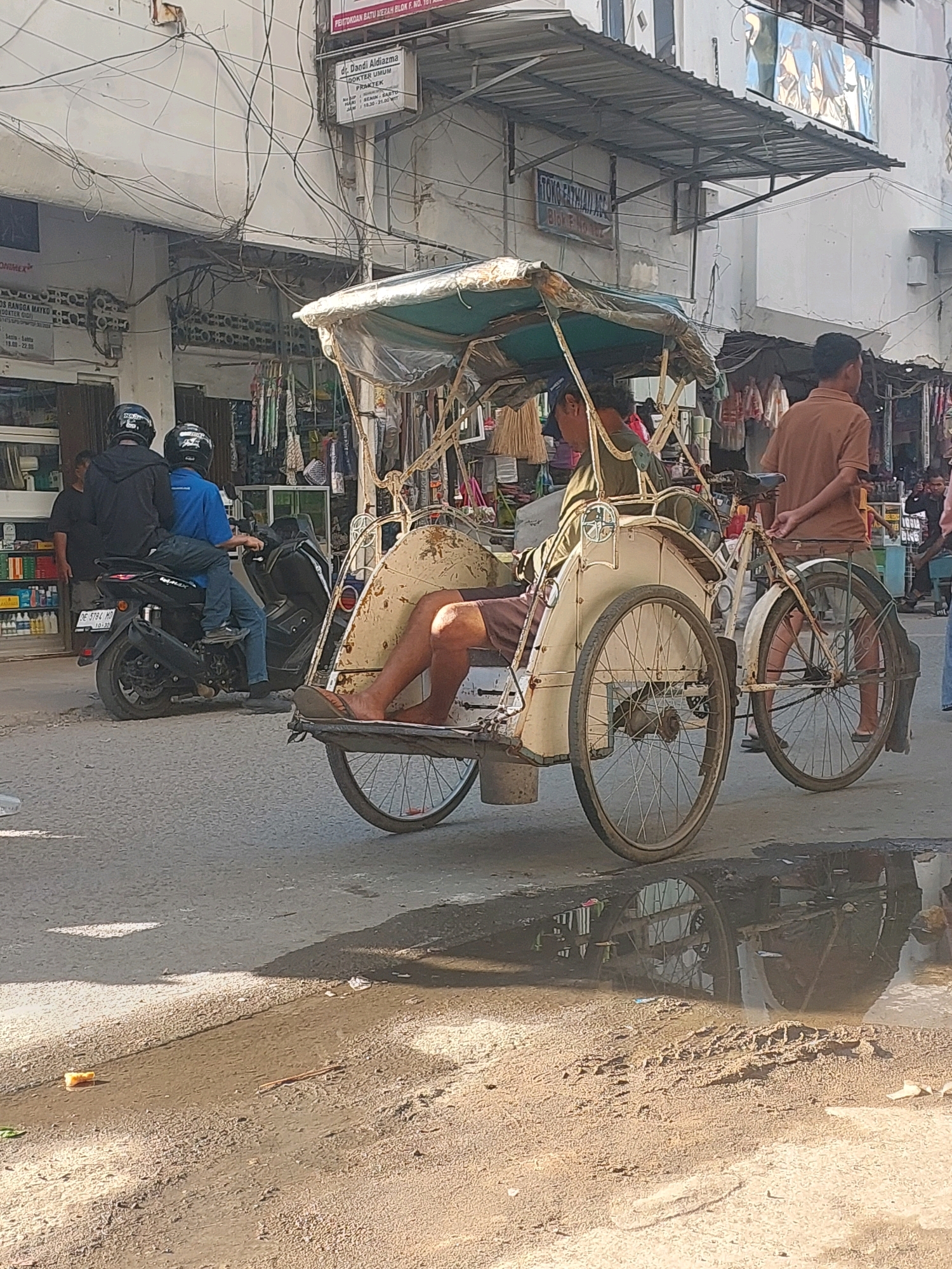 The image depicts a street scene in Ambon, Indonesia. In the foreground, a becak, a three-wheeled passenger vehicle, is parked. The becak is weathered and appears to be made of metal with a fabric canopy. A person's legs, wearing sandals, are visible in the passenger seat of the becak. Behind the becak, a man in a brown shirt stands with his hands behind his back. Further to the left, two men on a black motorcycle, both wearing helmets, are stopped. The motorcycle is a modern scooter. The environment is a street with buildings on either side. The buildings are two to three stories high and show signs of age and wear, with visible electrical wiring overhead. The street surface is asphalt, with patches of dirt and puddles, suggesting recent rain or a lack of proper drainage. The overall impression is a busy, somewhat unkempt urban environment.
