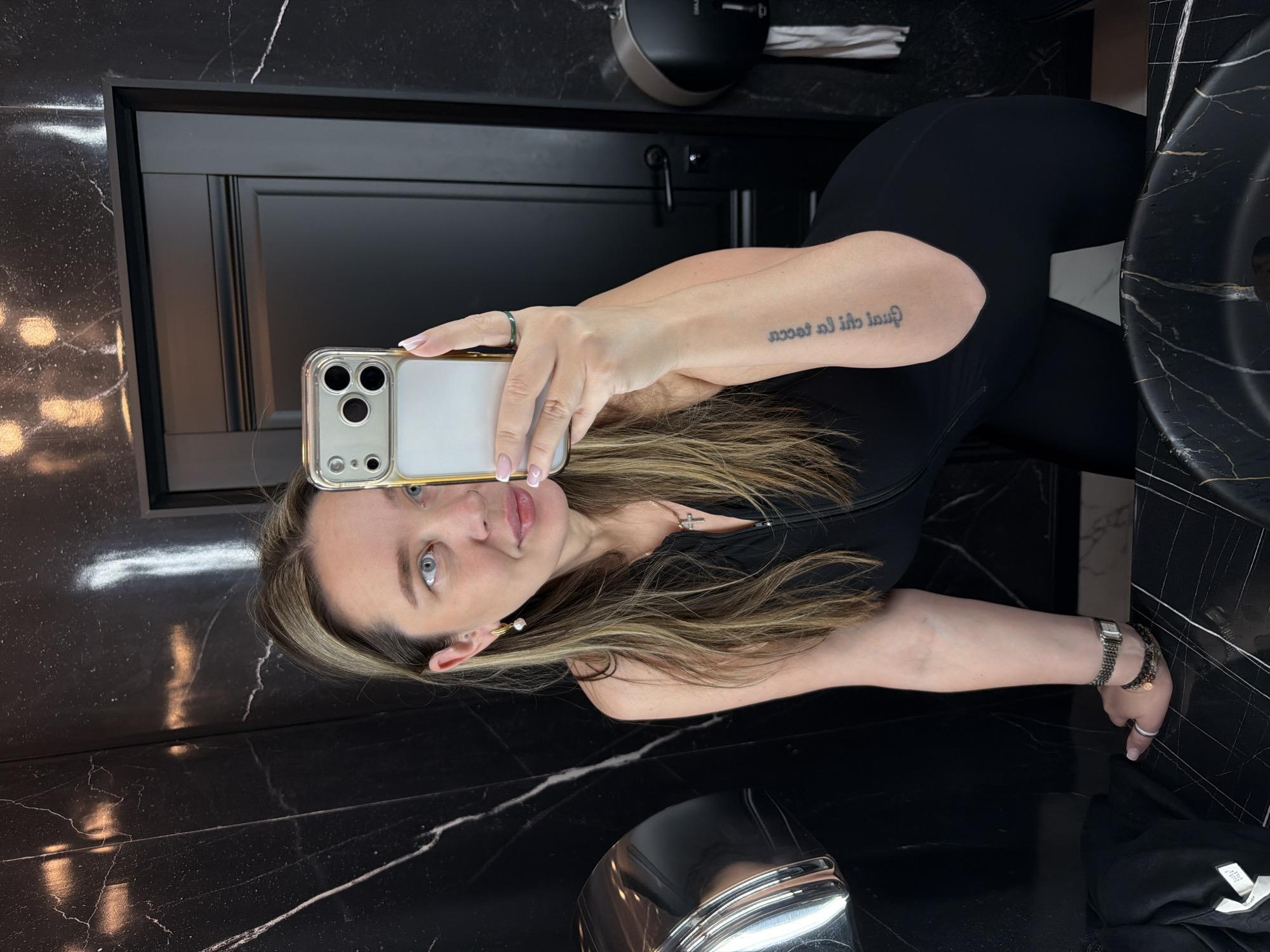 A fair-skinned woman with blonde highlighted hair is taking a selfie in a bathroom with black marble walls. She is wearing a black jumpsuit and jewelry, including a ring on her left hand, a watch, and bracelets on her right wrist. She holds a phone in her left hand. The bathroom features a black marble countertop with a sink, a hand dryer on the wall, and a closed black door. The location is unspecified but not necessarily Moscow, Russia.
