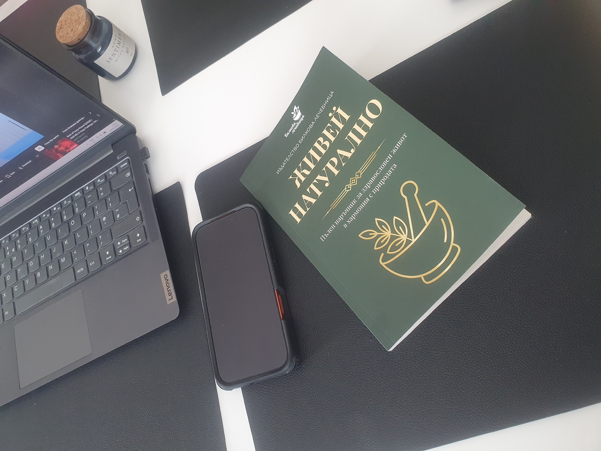 The image shows a desk with a black and white patterned surface. On the left, there is a grey Lenovo laptop with a video playing on the screen. Next to the laptop is a small jar labeled "HOME SENTIMENTS green beaver". In the center, there is a smartphone and a green book titled "ЖИВЕЙ НАТУРАЛНО" (Live Naturally) in Bulgarian, which is described as "Пълен наръчник за здравословен живот в хармония с природата" (A complete guide for a healthy life in harmony with nature) and is published by "ИЗДАТЕЛСТВО БИЛКОВА ЛЕЧЕБНИЦА" (Herbal Pharmacy Publishing). The image does not provide enough information to determine if the location is in Plovdiv, Bulgaria.