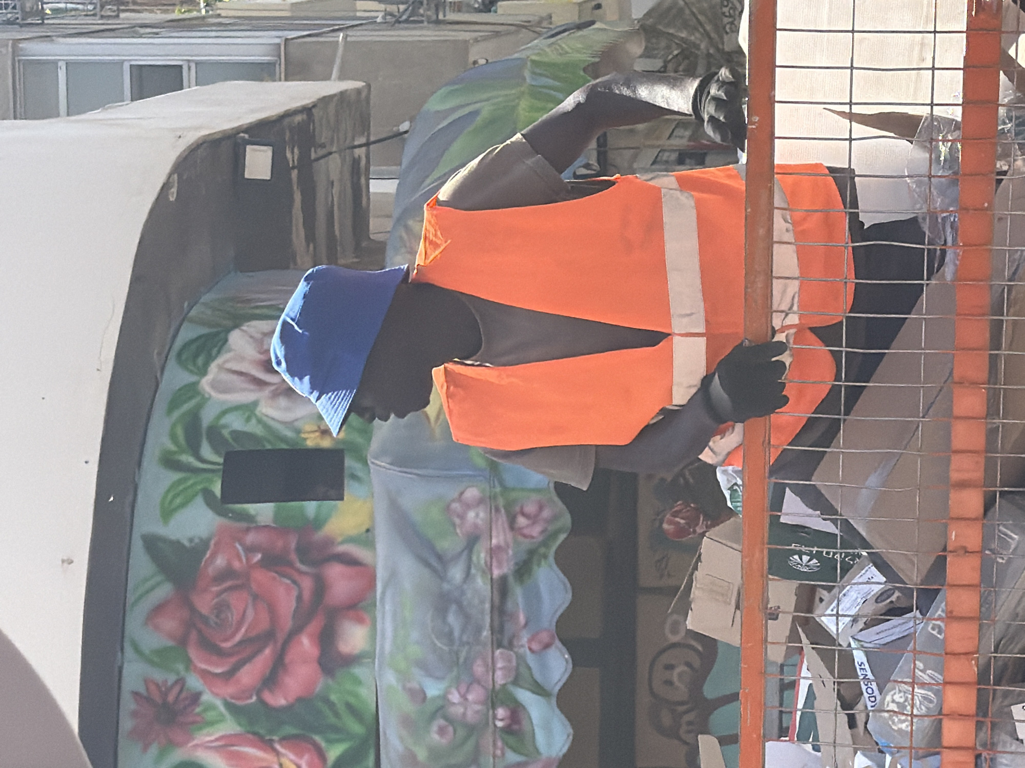 The image depicts a man in Tel-Aviv, Israel, wearing a blue cap, orange safety vest, and black gloves. He appears to be behind a metal cage, possibly involved in waste collection or recycling, as indicated by the cardboard boxes inside. The man is positioned next to a painted dumpster decorated with floral designs, including a prominent red rose. The background includes other buildings, suggesting an urban environment. The scene captures a moment of labor in an everyday urban setting.