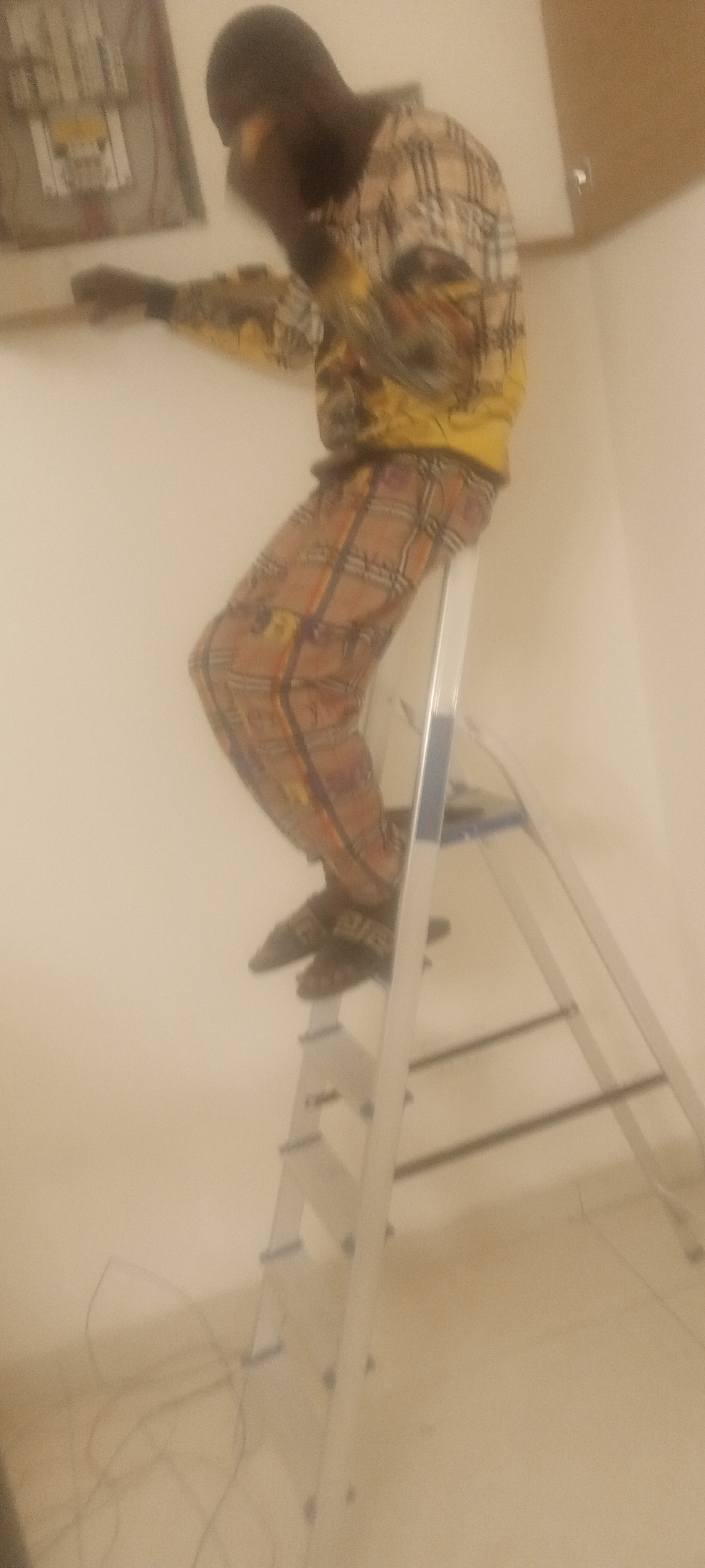 A blurred image shows a dark-skinned person in patterned clothing standing on a silver stepladder in what appears to be an indoor setting, possibly in a building in Unguwar Rimi, Nigeria. The person is reaching towards a panel on the wall. Electrical wires are visible near the base of the ladder.